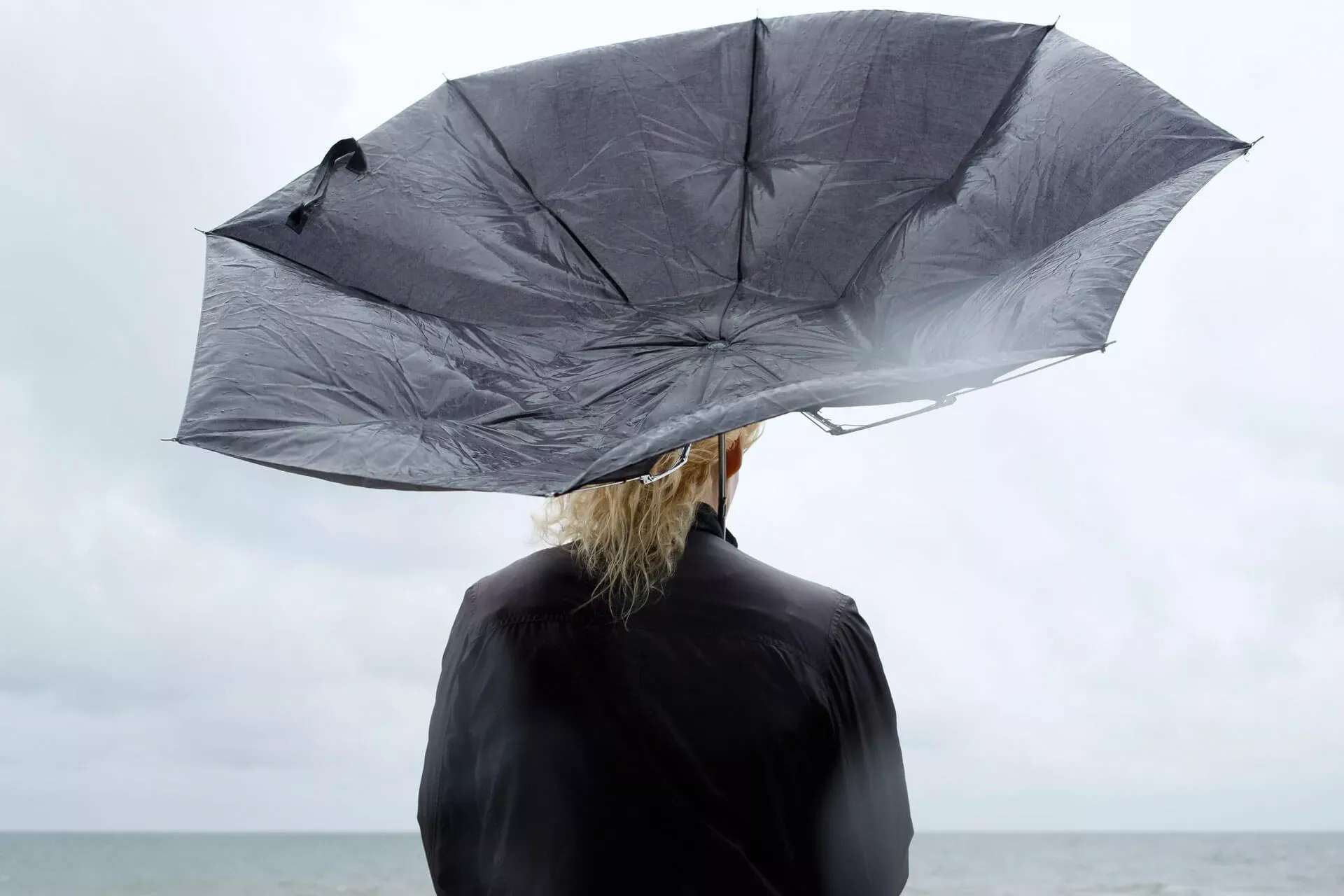 Frau steht mit schwarzem Schirm in Regensturm und schaut auf das Meer.– Bild: Folio Images RF © Strandperle Frau steht mit schwarzem Schirm in Regensturm und schaut auf das Meer.– Bild: Folio Images RF © Strandperle
