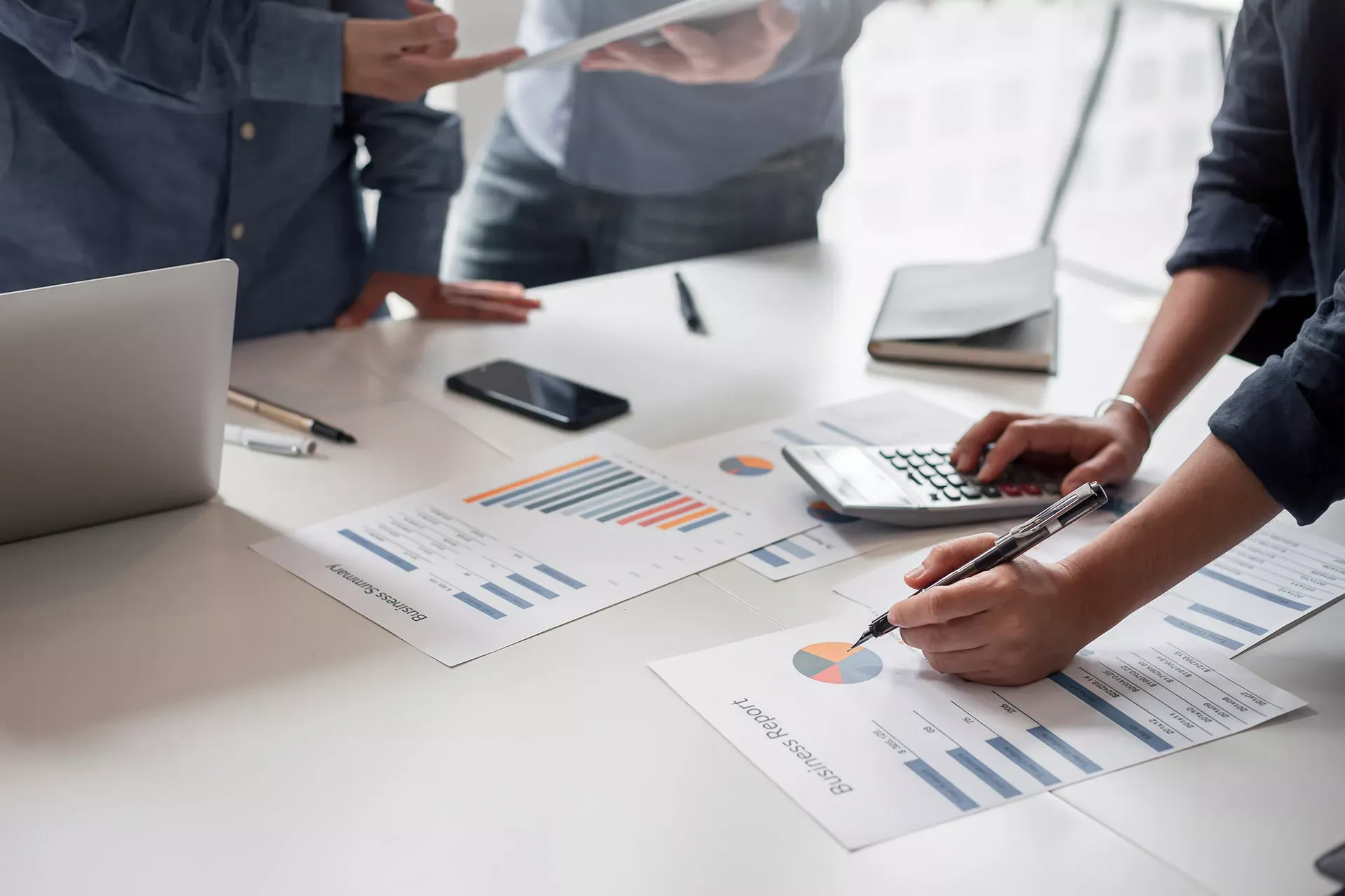 Gruppe von Menschen stehen um einen Tisch mit Finanzcharts, Taschenrechner, Laptop, Smartphone und Stift. Gruppe von Menschen stehen um einen Tisch mit Finanzcharts, Taschenrechner, Laptop, Smartphone und Stift.