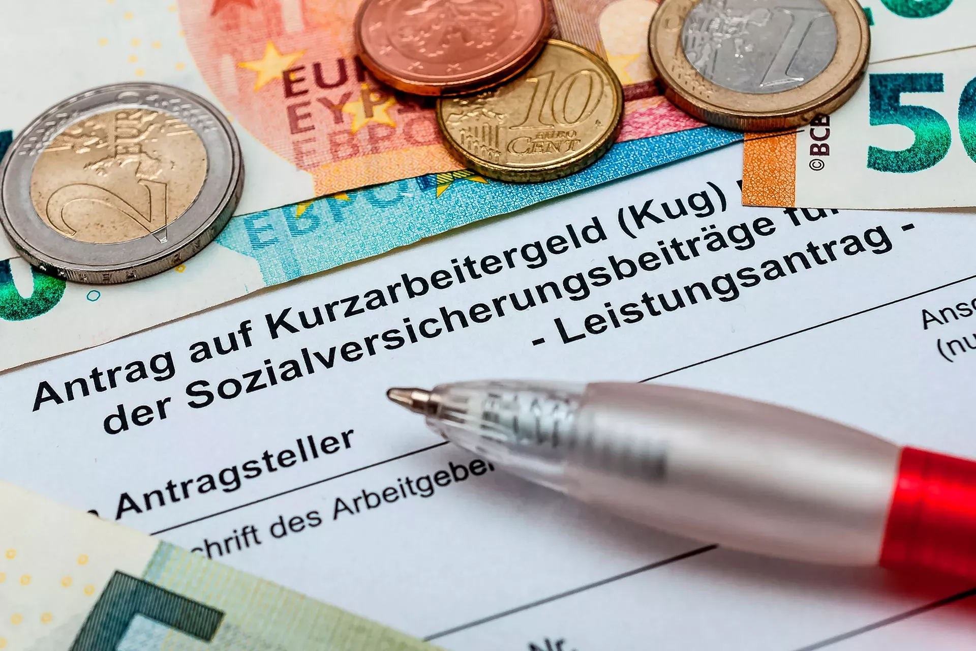 Antrag auf Kurzarbeitergeld, darauf liegen ein Kugelschreiber sowie Münzen und Geldscheine. Antrag auf Kurzarbeitergeld, darauf liegen ein Kugelschreiber sowie Münzen und Geldscheine.