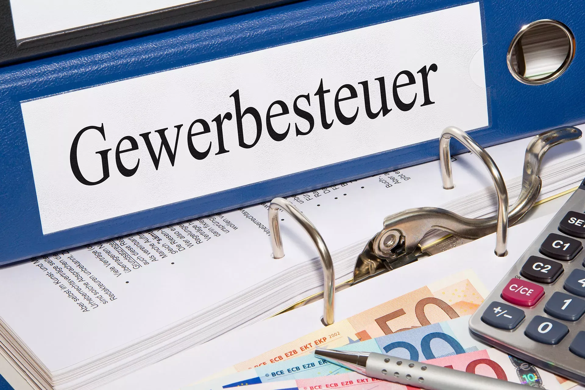 Ordner mit der Aufschrift Gewerbesteuer liegt auf einem geöffneten Ordner, daneben Geldscheine, Kugelschreiber und ein Taschenrechner. Ordner mit der Aufschrift Gewerbesteuer liegt auf einem geöffneten Ordner, daneben Geldscheine, Kugelschreiber und ein Taschenrechner.