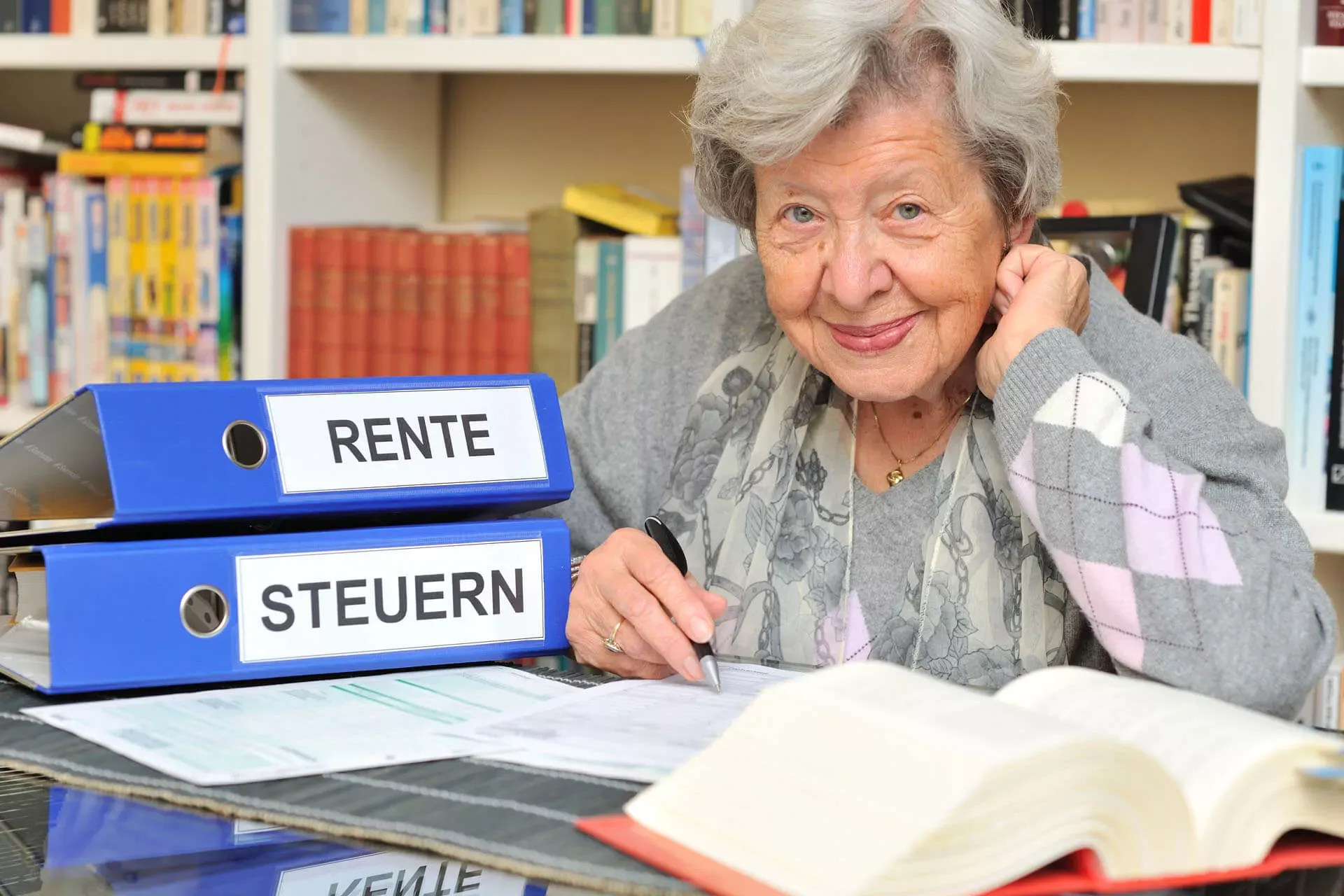 Rentnerin sitzt an einem Tisch mit Ordnern Steuer und Rente sowie einem Steuergesetzbuch und füllt ein Steuererklärungsformular aus. Rentnerin sitzt an einem Tisch mit Ordnern Steuer und Rente sowie einem Steuergesetzbuch und füllt ein Steuererklärungsformular aus.