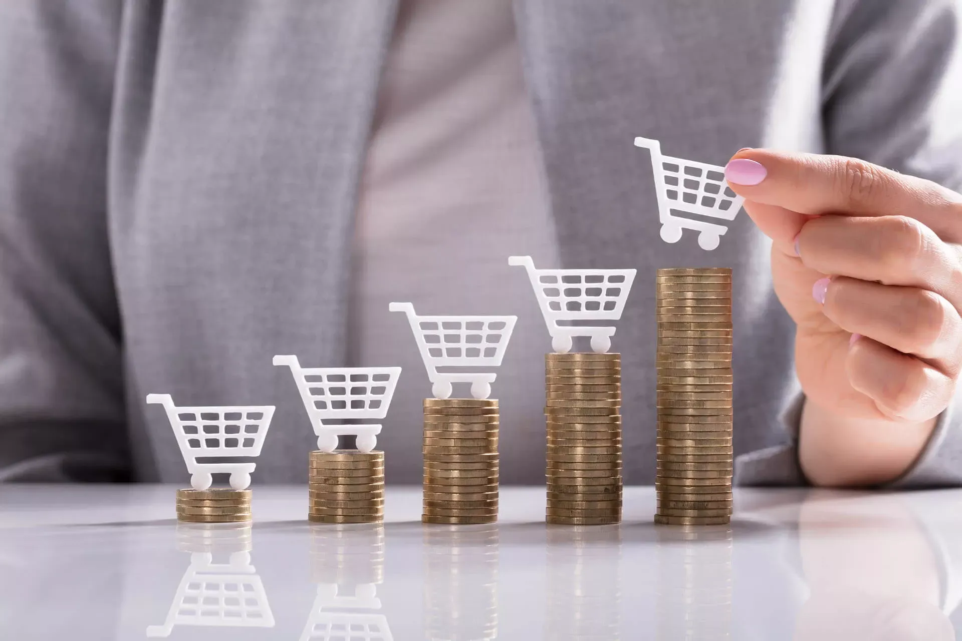 Frau in grauem Kostüm hält ein weißes Einkaufswagen-Modell über einem Stapel Münzen in die Höhe, links daneben weitere, niedriger werdende Stapel Münzen mit jeweils einem Einkaufswagen darauf. Frau in grauem Kostüm hält ein weißes Einkaufswagen-Modell über einem Stapel Münzen in die Höhe, links daneben weitere, niedriger werdende Stapel Münzen mit jeweils einem Einkaufswagen darauf.