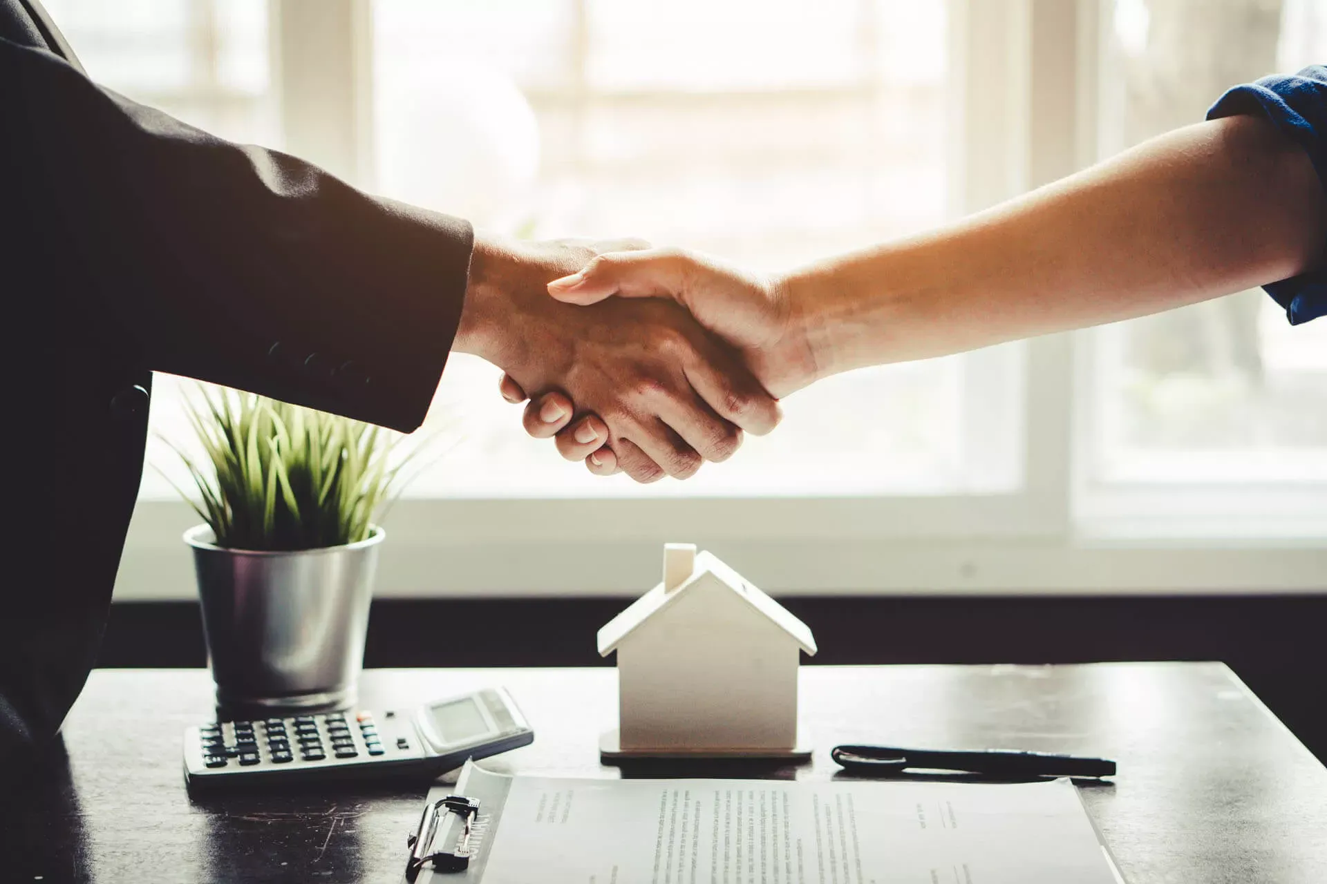 Immobilienfinanzierung: Händeschütteln über einem Tisch mit einem Hausmodell, einem Vertrag und einem Stift. Immobilienfinanzierung: Händeschütteln über einem Tisch mit einem Hausmodell, einem Vertrag und einem Stift.