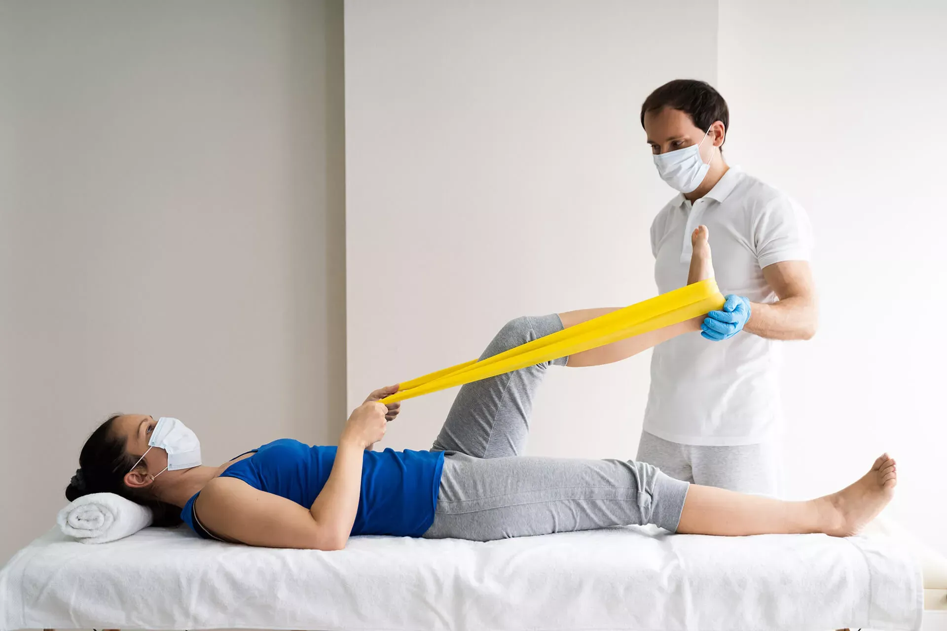 Physiotherapeut hilft Reha-Patientin bei einer Übung für das Knie mit einem gelben Theraband. Beide tragen eine Corona-Schutzmaske. Physiotherapeut hilft Reha-Patientin bei einer Übung für das Knie mit einem gelben Theraband. Beide tragen eine Corona-Schutzmaske.