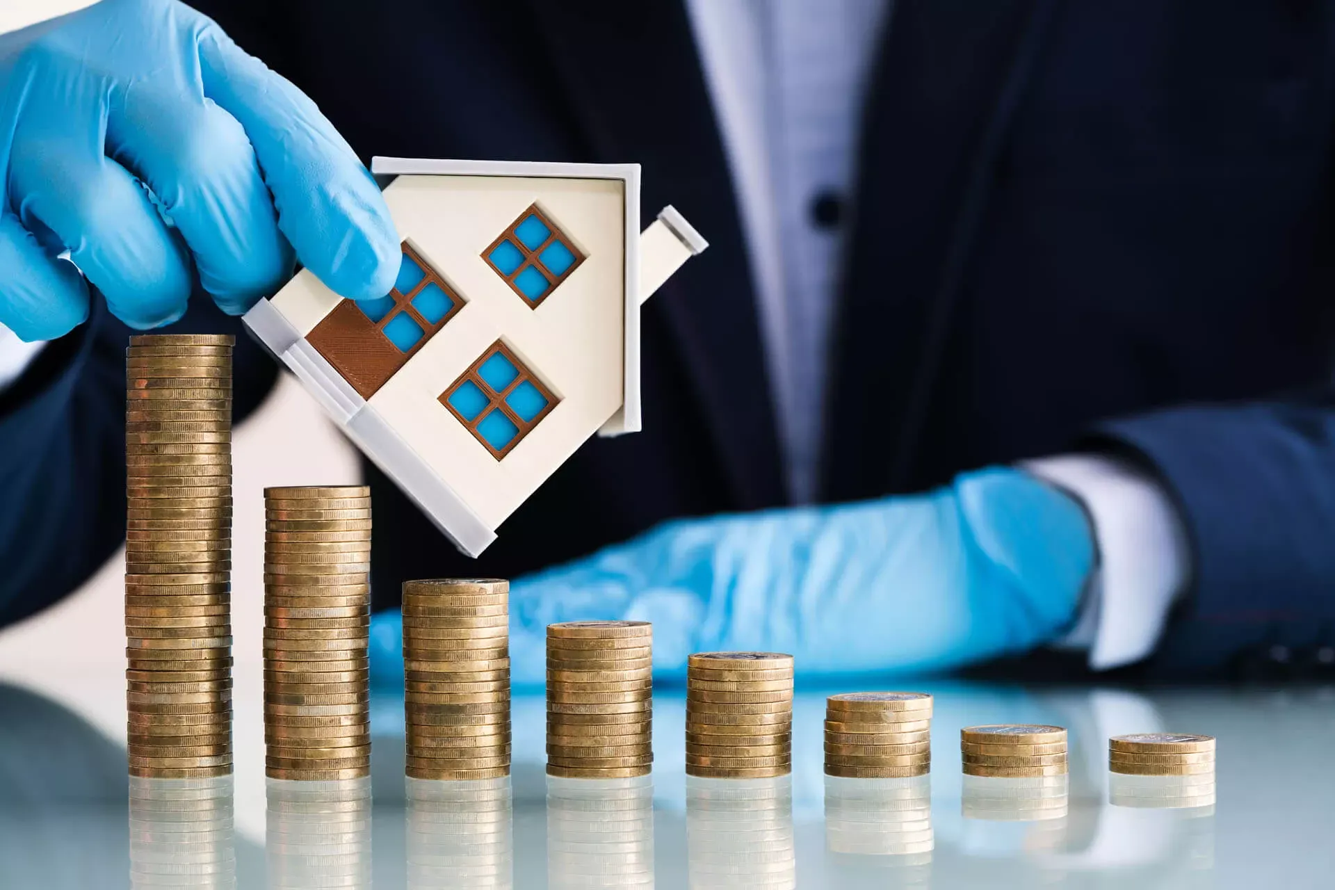 Immobilienpreise in Corona-Zeiten: Mann im Anzug trägt blaue Handschuhe und hält ein Hausmodell schräg über eine Reihe von Münzstapeln, die von links nach rechts niedriger werden. Immobilienpreise in Corona-Zeiten: Mann im Anzug trägt blaue Handschuhe und hält ein Hausmodell schräg über eine Reihe von Münzstapeln, die von links nach rechts niedriger werden.