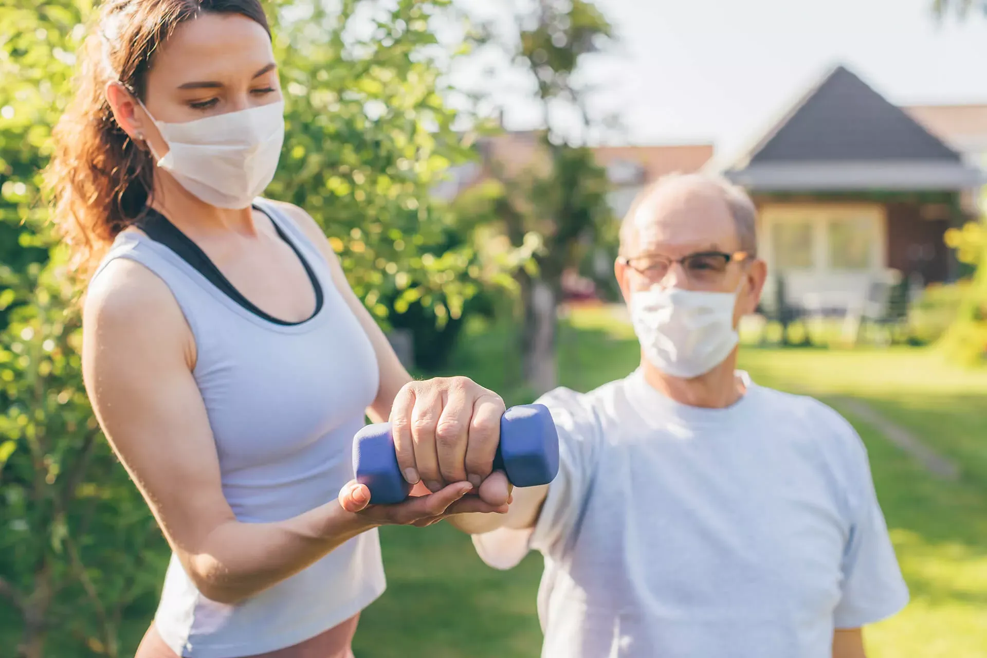 Physiotherapeutin hilft älterem Reha-Patient beim Hanteltraining im Freien, beide tragen eine Corona-Schutzmaske. Physiotherapeutin hilft älterem Reha-Patient beim Hanteltraining im Freien, beide tragen eine Corona-Schutzmaske.