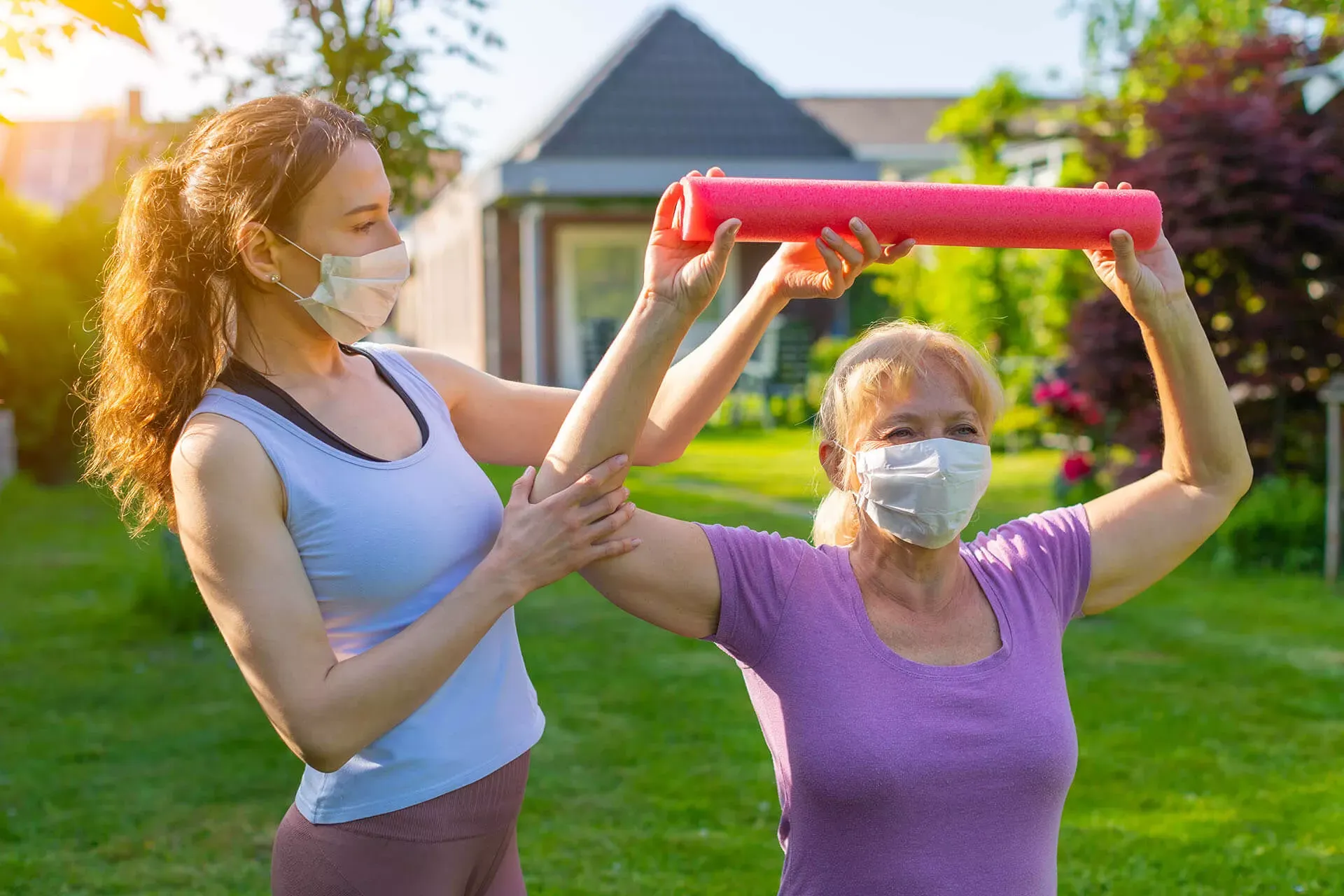 Physiotherapeutin steht mit älterer Reha-Patientin auf einer Wiese und unterstützt sie bei Theraband-Übungen. Beide tragen einen Mund-Nasen-Schutz. Physiotherapeutin steht mit älterer Reha-Patientin auf einer Wiese und unterstützt sie bei Theraband-Übungen. Beide tragen einen Mund-Nasen-Schutz.