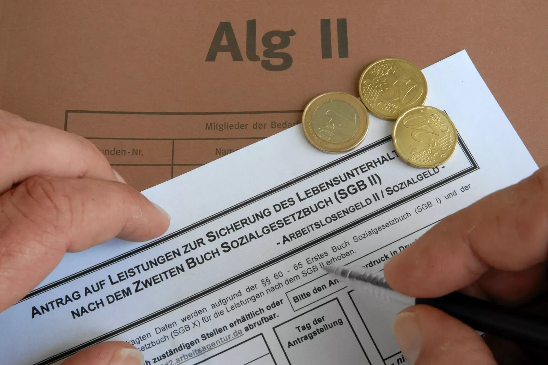 Hand füllt ALG-2-Antrag aus, dabei liegen Geldmünzen und ALG-2-Umschlag. Hand füllt ALG-2-Antrag aus, dabei liegen Geldmünzen und ALG-2-Umschlag.