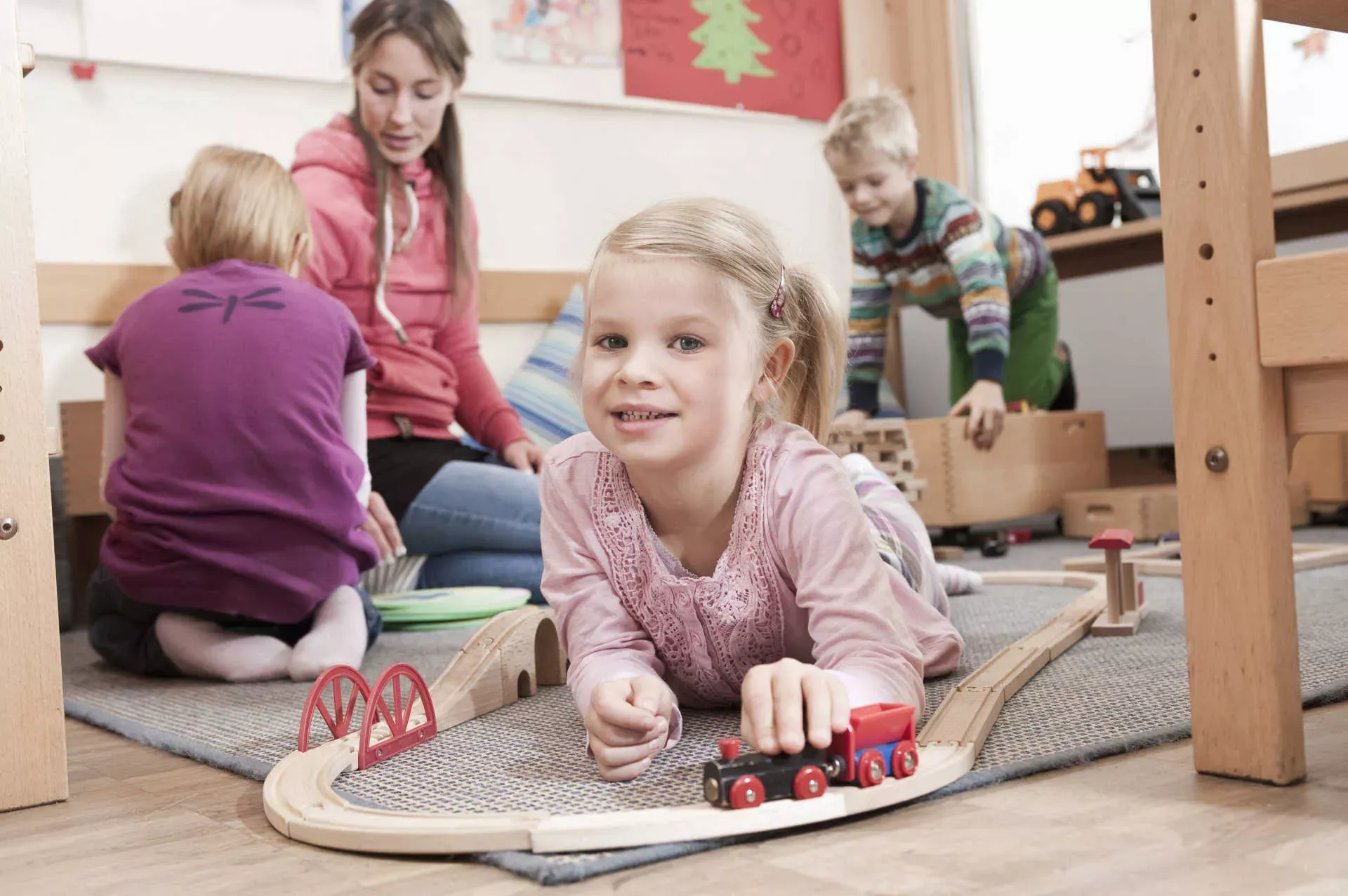 Mädchen in Kita spielt mit Holzzug auf dem Boden, im Hintergrung Erzieherin und weitere Kinder. Bild: IMAGO / Cavan Images/ imago stock&people Mädchen in Kita spielt mit Holzzug auf dem Boden, im Hintergrung Erzieherin und weitere Kinder. Bild: IMAGO / Cavan Images/ imago stock&people