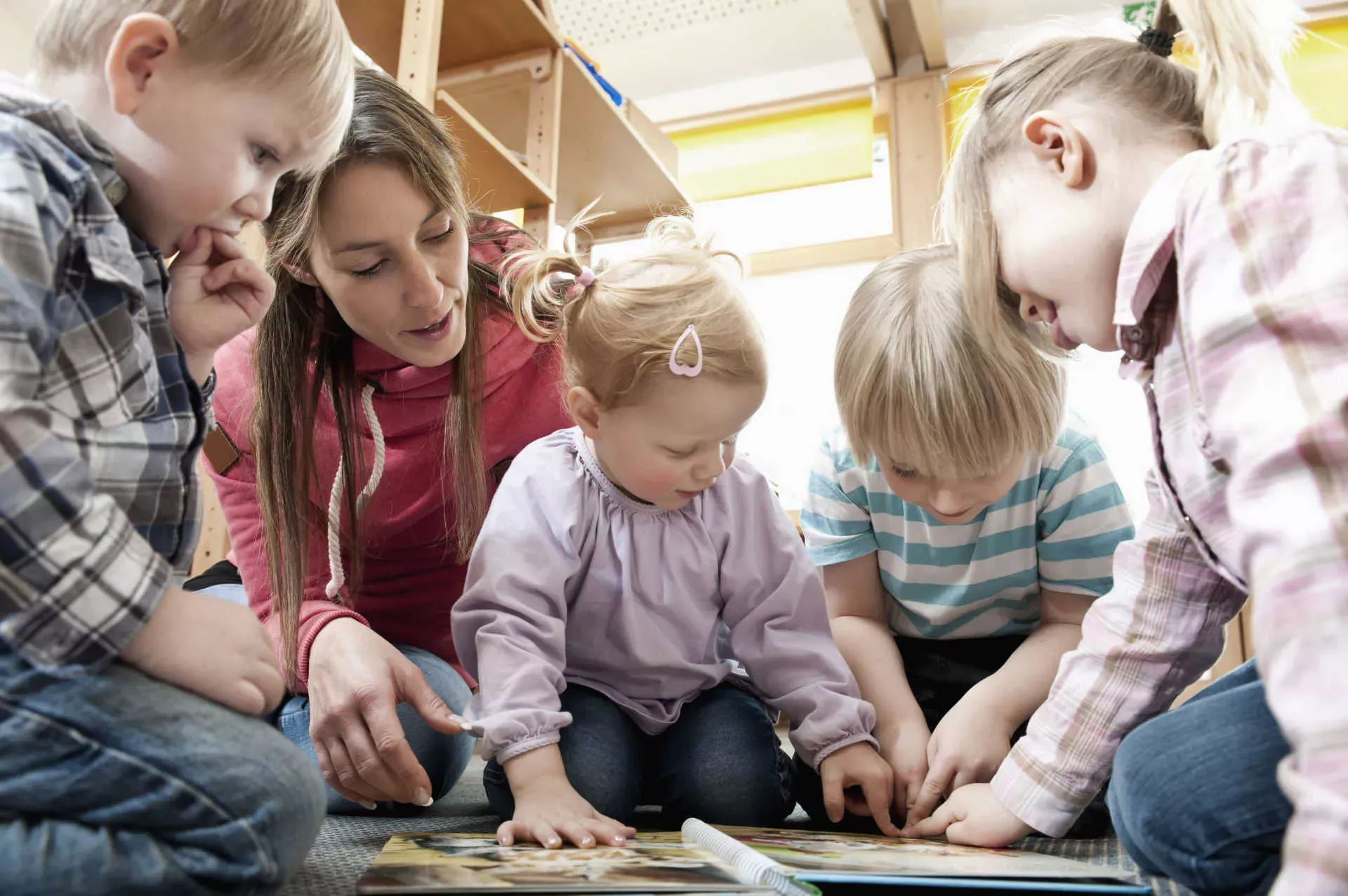 Erzieherin in Kita schaut mit kleinen Kindern ein Bilderbuch auf dem Boden an. Bild: IMAGO / Cavan Images / imago stock&people Erzieherin in Kita schaut mit kleinen Kindern ein Bilderbuch auf dem Boden an. Bild: IMAGO / Cavan Images / imago stock&people