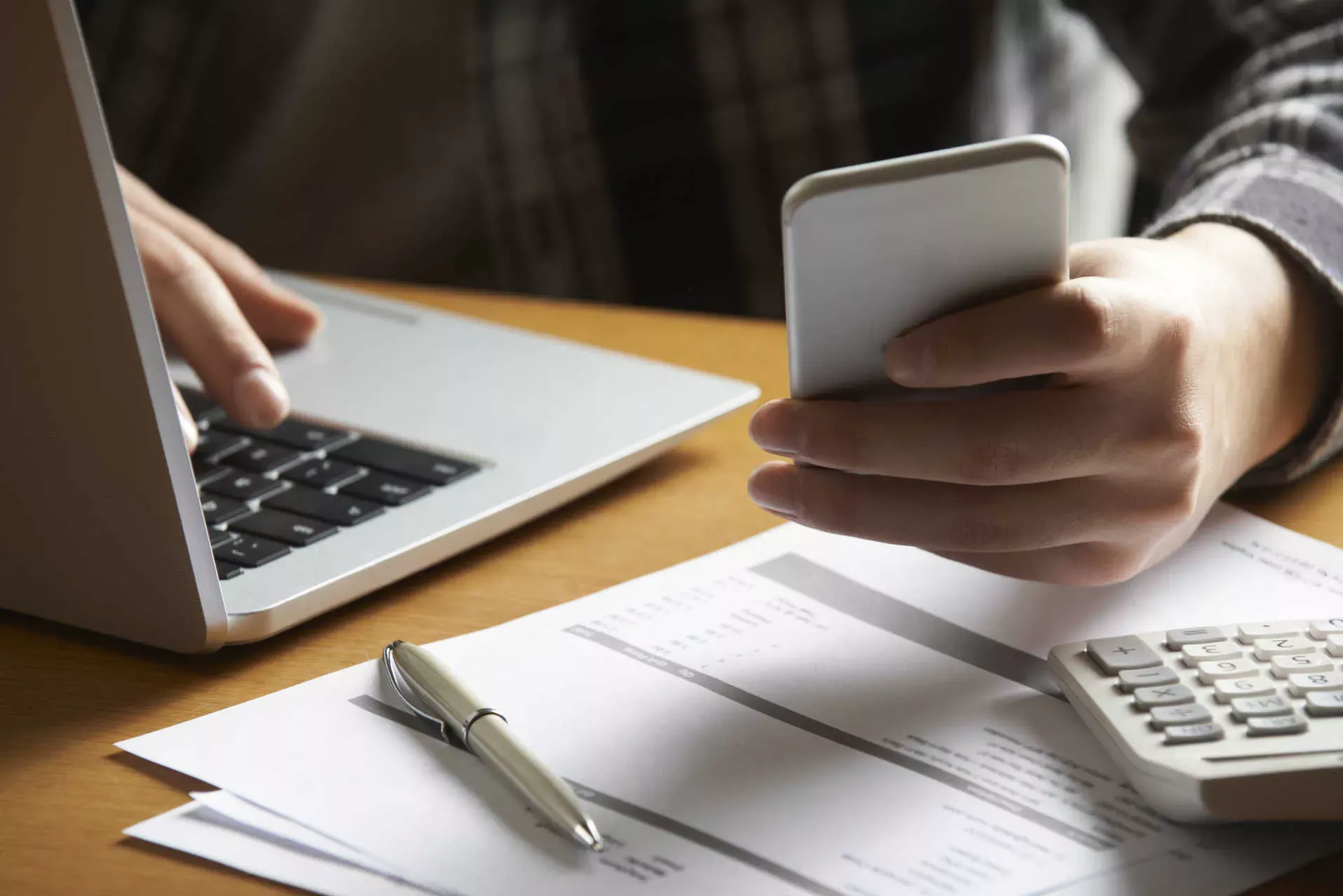Frau bedient Smartphone und Notebook, daneben Unterlagen, Taschenrechner und Stift. Bild: IMAGO / ingimage Frau bedient Smartphone und Notebook, daneben Unterlagen, Taschenrechner und Stift. Bild: IMAGO / ingimage