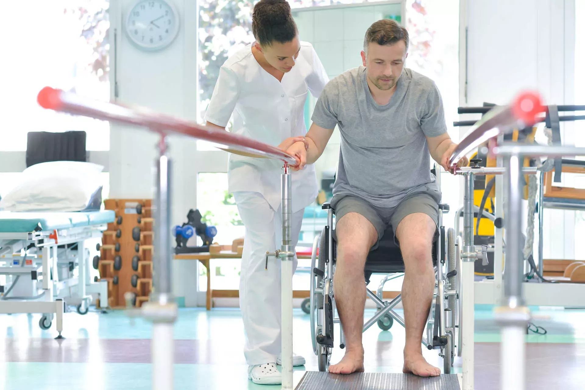 Physiotherapeutin hilft Reha-Patienten beim Aufstehen aus dem Rollstuhl, um Gehübungen am Barren zu machen. Bild: IMAGO / agefotostock / photography33 Physiotherapeutin hilft Reha-Patienten beim Aufstehen aus dem Rollstuhl, um Gehübungen am Barren zu machen. Bild: IMAGO / agefotostock / photography33