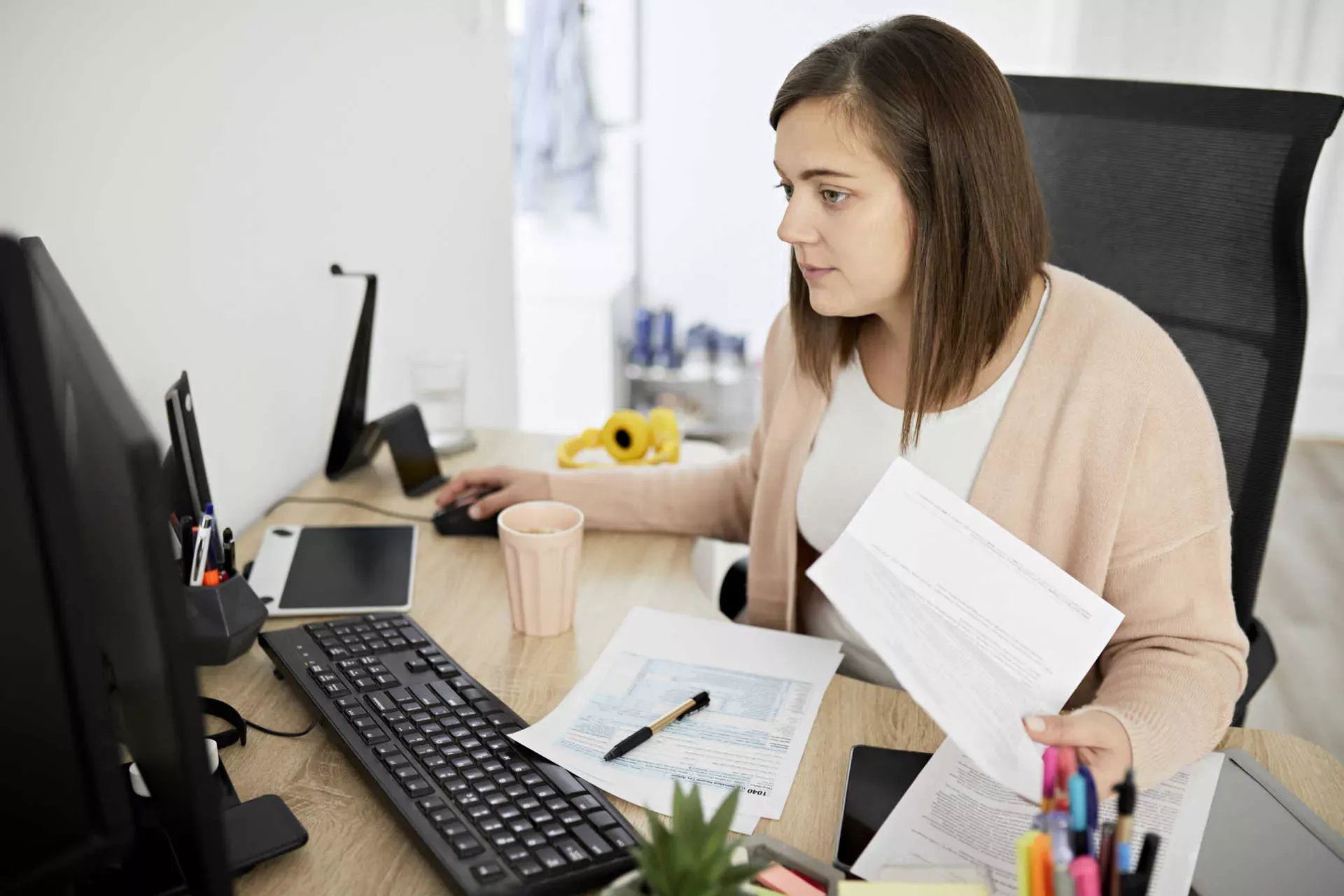 Junge Frau mit Unterlagen in der Hand bedient Computermaus. Bild: IMAGO / Westend61 / Zeljiko Dangubic Junge Frau mit Unterlagen in der Hand bedient Computermaus. Bild: IMAGO / Westend61 / Zeljiko Dangubic