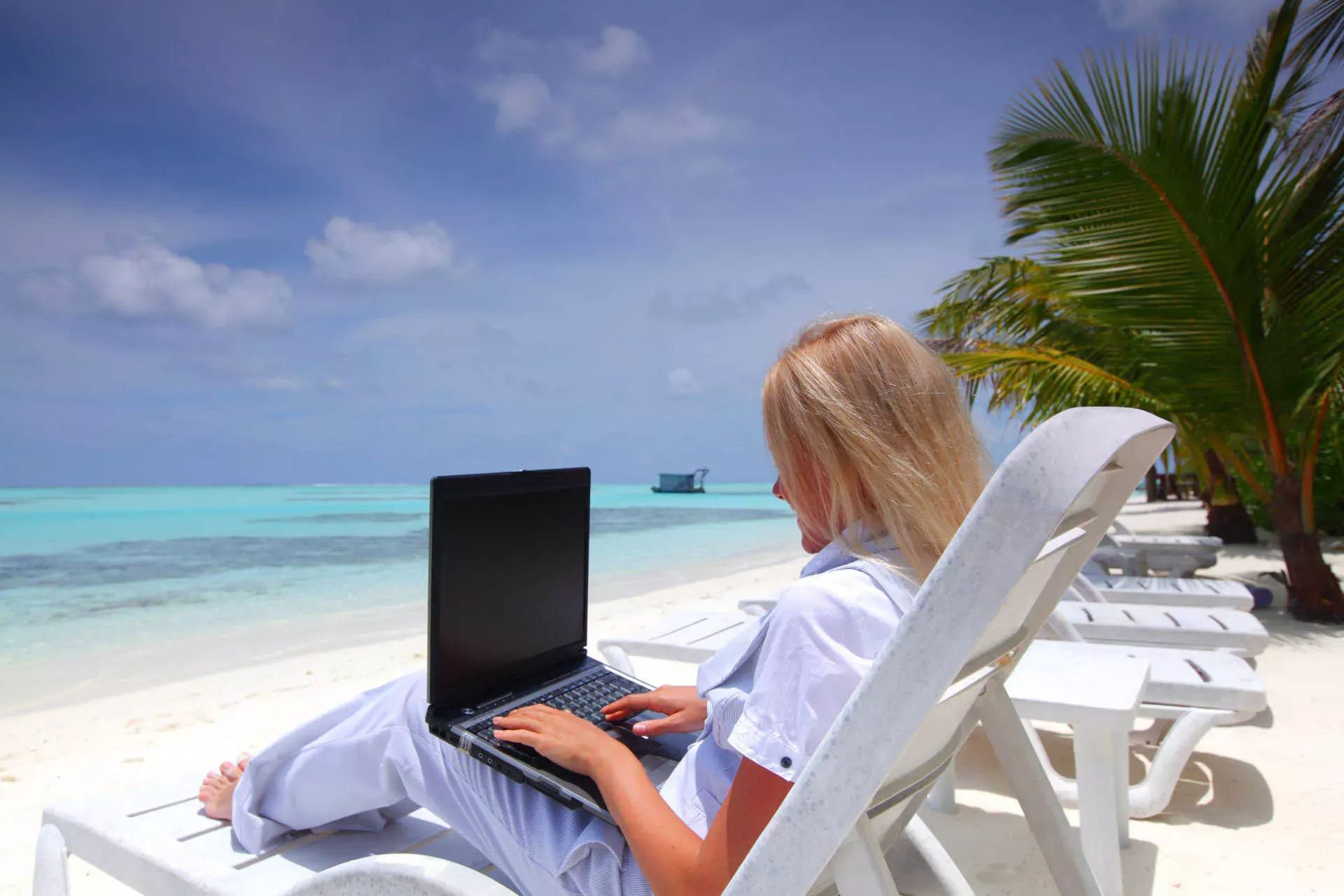 Junge blonde Frau in Businesskleidung sitzt an einem Strand in einem Liegestuhl und arbeitet an ihrem Notebook. Bild: IMAGO / YAY Images Junge blonde Frau in Businesskleidung sitzt an einem Strand in einem Liegestuhl und arbeitet an ihrem Notebook. Bild: IMAGO / YAY Images