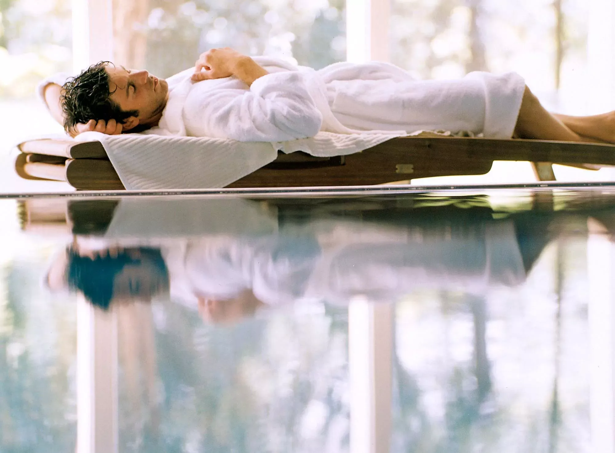 Junger Mann im Bademantel liegt am Rand eines Wellness-Beckens auf einer Liege und entspannt sich. Junger Mann im Bademantel liegt am Rand eines Wellness-Beckens auf einer Liege und entspannt sich.