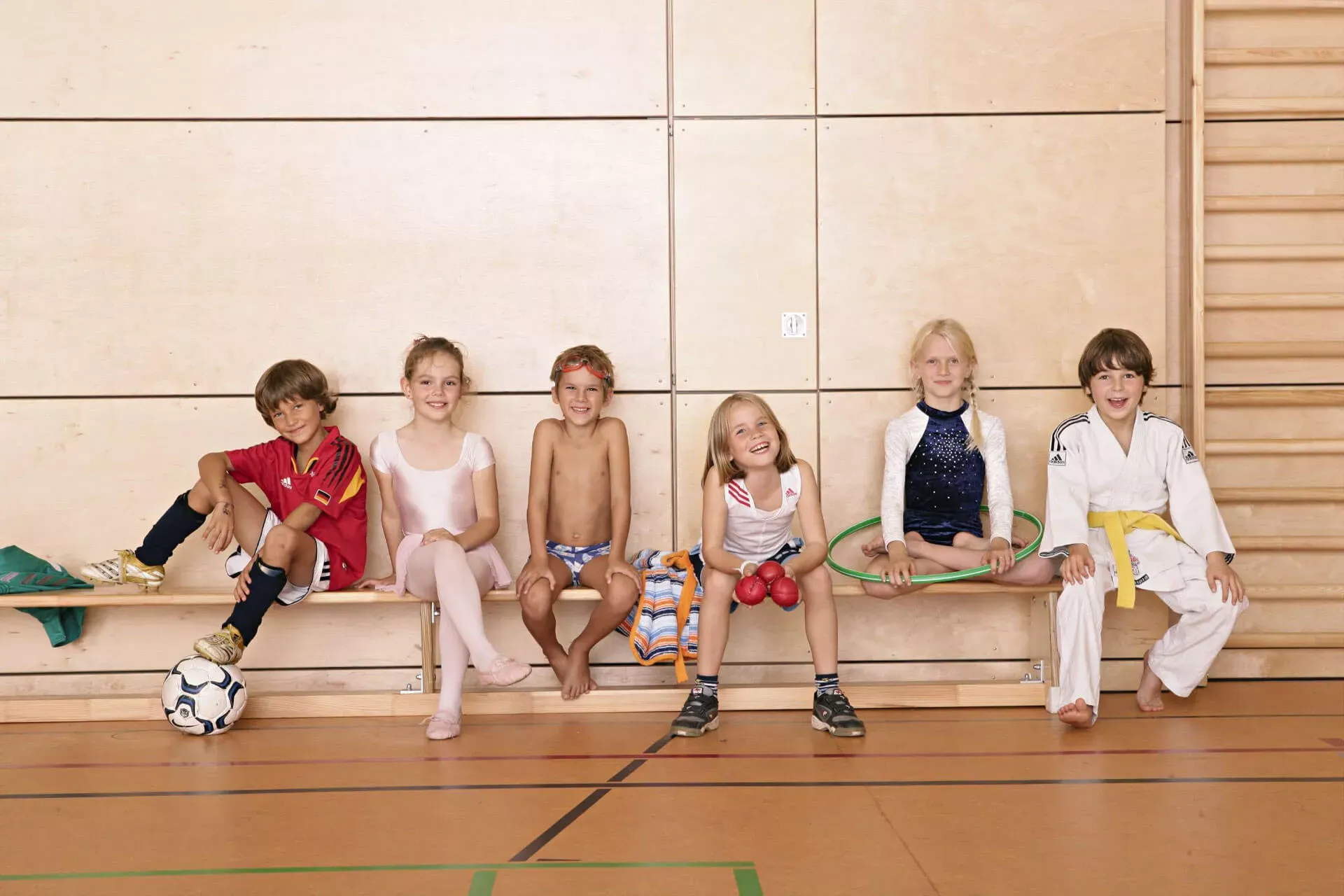 Eine Gruppe Kinder in Sportkleidung unterschiedlicher Sportarten sitzt auf einer Bank in einer Turnhalle. Eine Gruppe Kinder in Sportkleidung unterschiedlicher Sportarten sitzt auf einer Bank in einer Turnhalle.