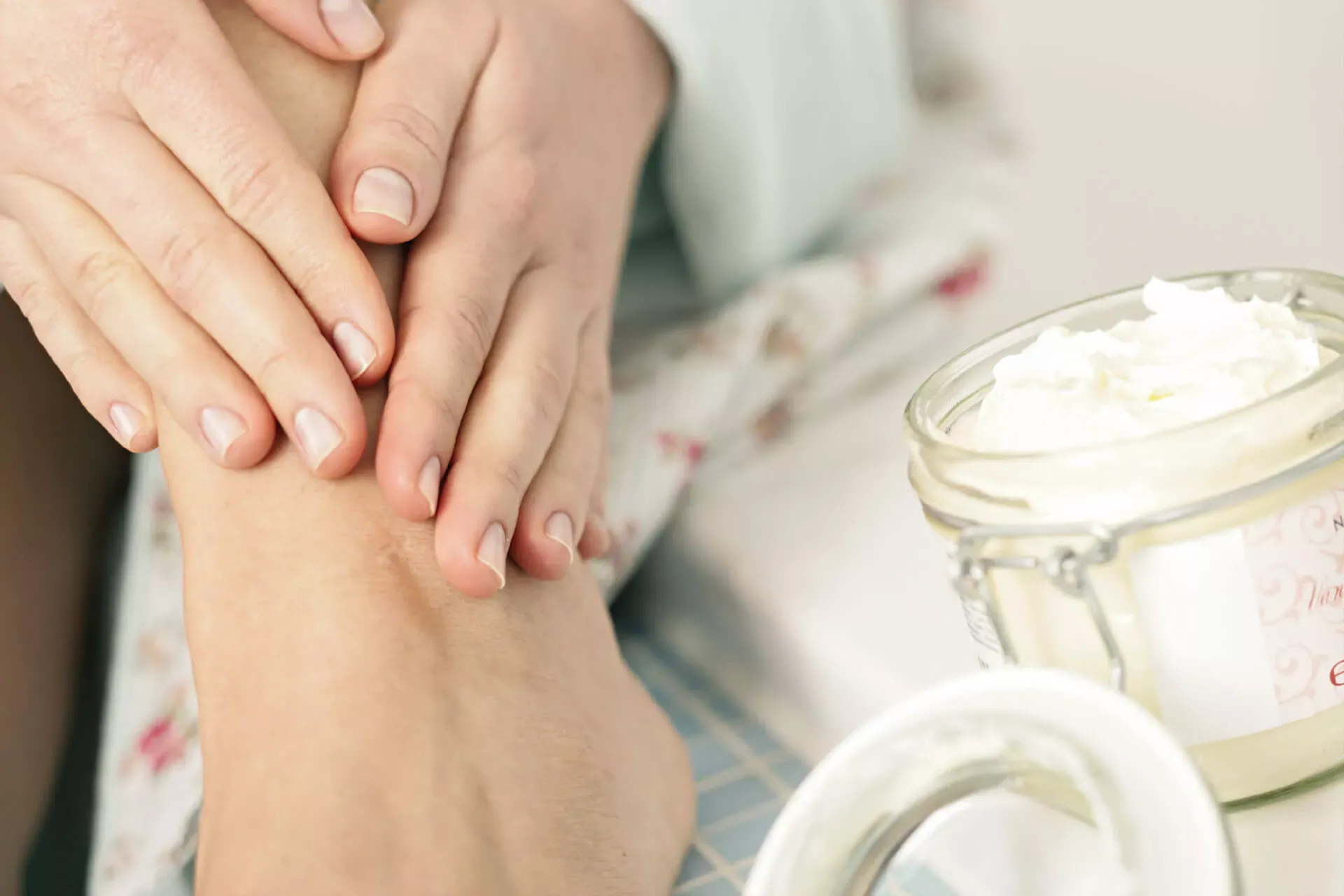 Frau cremt ihren Fuß ein, danben ein Glastopf mit weißer Creme. Frau cremt ihren Fuß ein, danben ein Glastopf mit weißer Creme.