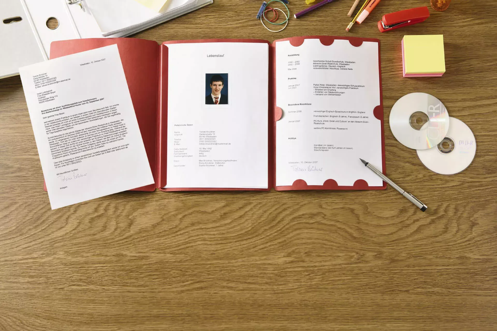Rote Bewerbungsmappe aufgeschlagen mit Lebenslauf und Bewerbungsschreiben, daneben Stift und CDs. Rote Bewerbungsmappe aufgeschlagen mit Lebenslauf und Bewerbungsschreiben, daneben Stift und CDs.