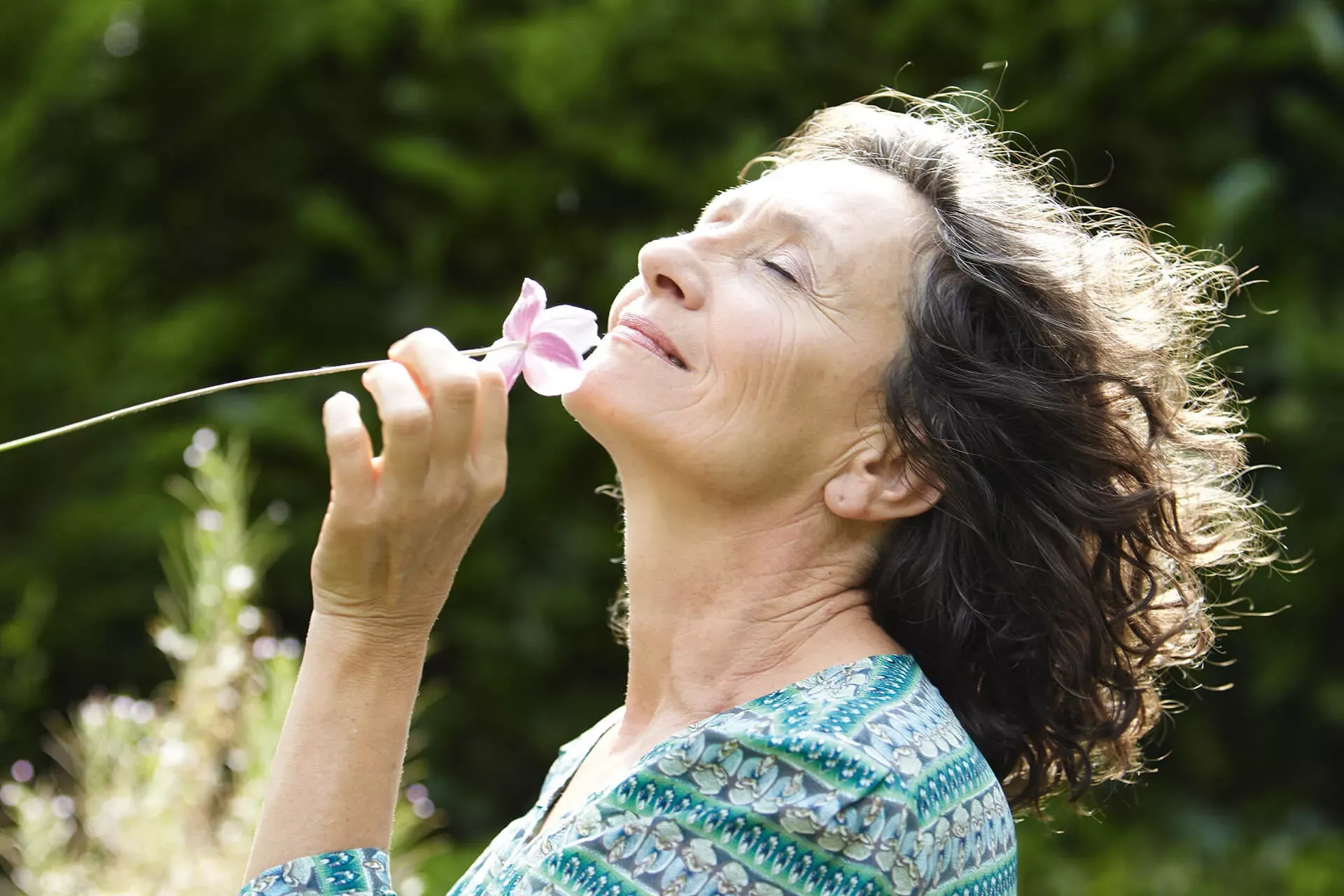 Ältere Frau steht in der Natur und atmet mit geschlossenen Augen den Duft einer Blume ein. Ältere Frau steht in der Natur und atmet mit geschlossenen Augen den Duft einer Blume ein.