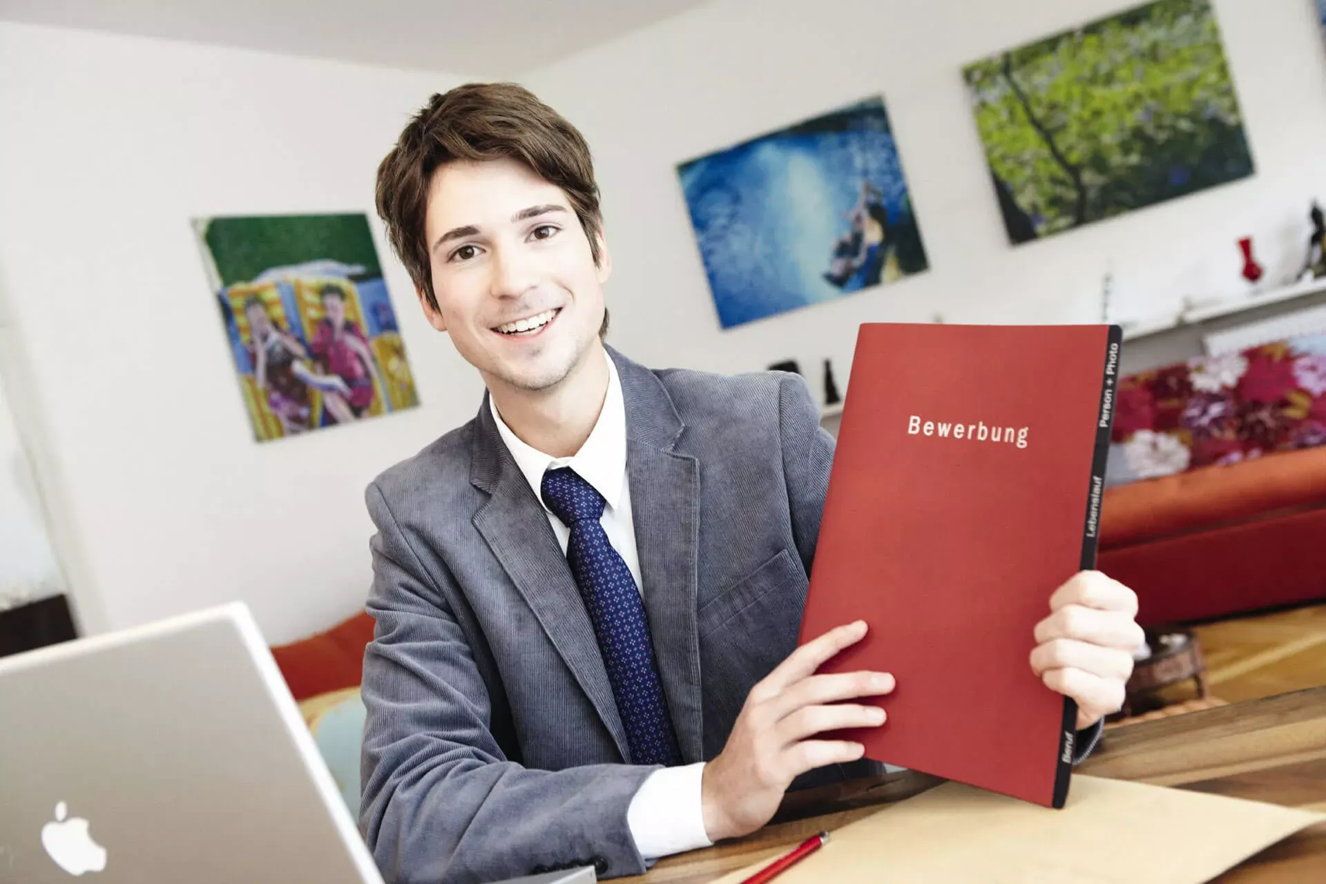 Junger Mann im Anzug sitzt an seinem Schreibtisch zuhause und hält eine Bewerbungsmappe. Junger Mann im Anzug sitzt an seinem Schreibtisch zuhause und hält eine Bewerbungsmappe.