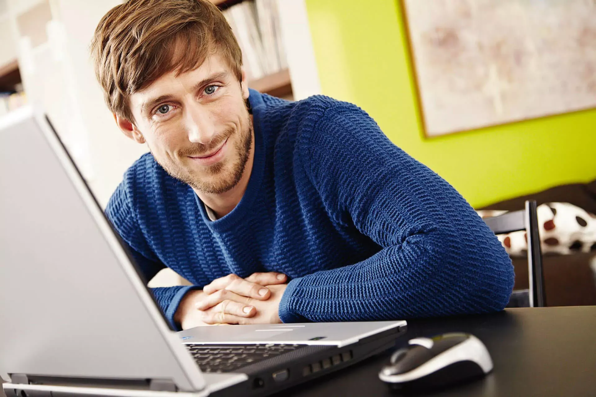 Junger Mann sitzt am Schreibtisch im Homeoffice vor einem Notebook und lächelt in die Kamera. Junger Mann sitzt am Schreibtisch im Homeoffice vor einem Notebook und lächelt in die Kamera.