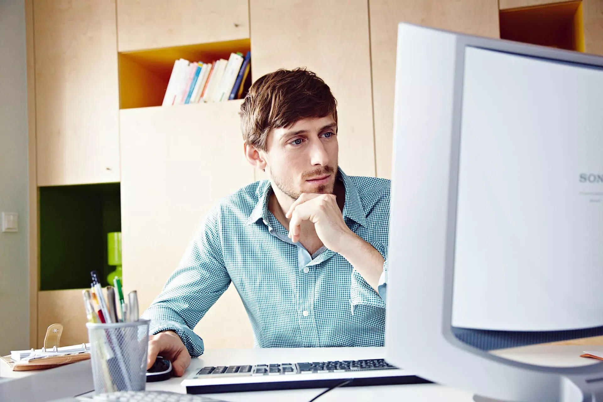 Junger Mann im Homeoffice sitzt in kariertem Hemd am Schreibtisch und schaut nachdenklich auf einen Computer-Bildschirm, der schräg vor ihm steht.. Junger Mann im Homeoffice sitzt in kariertem Hemd am Schreibtisch und schaut nachdenklich auf einen Computer-Bildschirm, der schräg vor ihm steht..