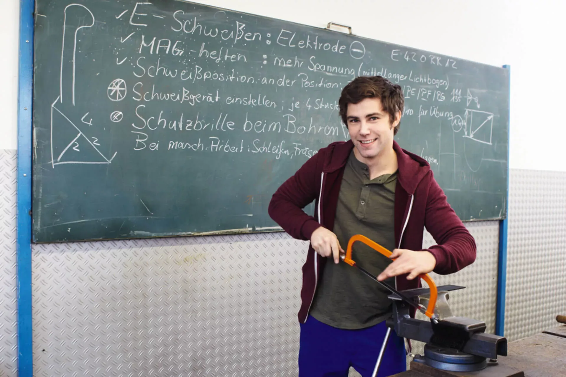 Männlicher Azubi steht in der Berufsschule vor einer Tafel und sägt an einem Metallwerkstück. Männlicher Azubi steht in der Berufsschule vor einer Tafel und sägt an einem Metallwerkstück.