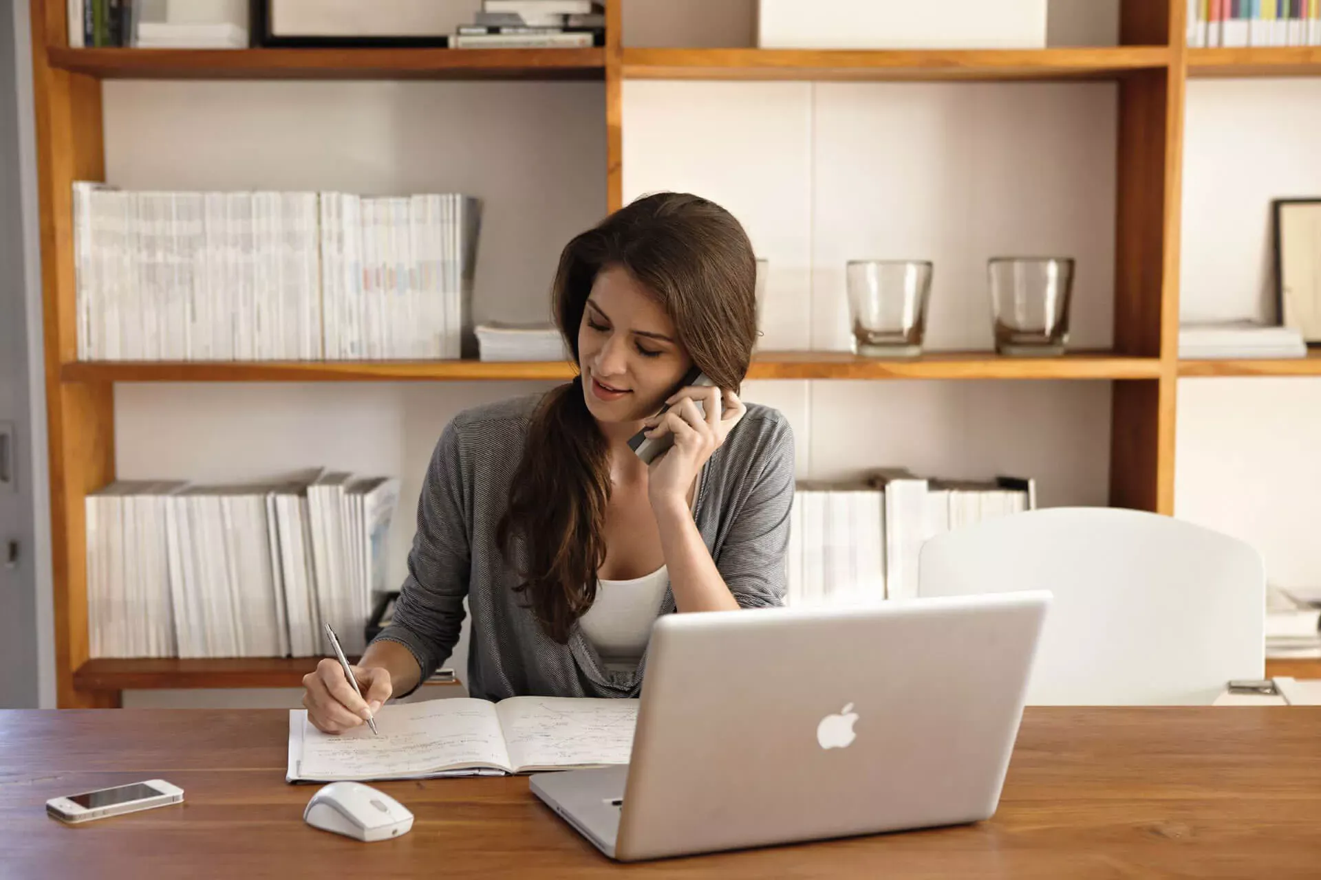 Junge Frau sitzt im Homoffice am Notebook, telefoniert und macht Notizen in ein Heft. Junge Frau sitzt im Homoffice am Notebook, telefoniert und macht Notizen in ein Heft.