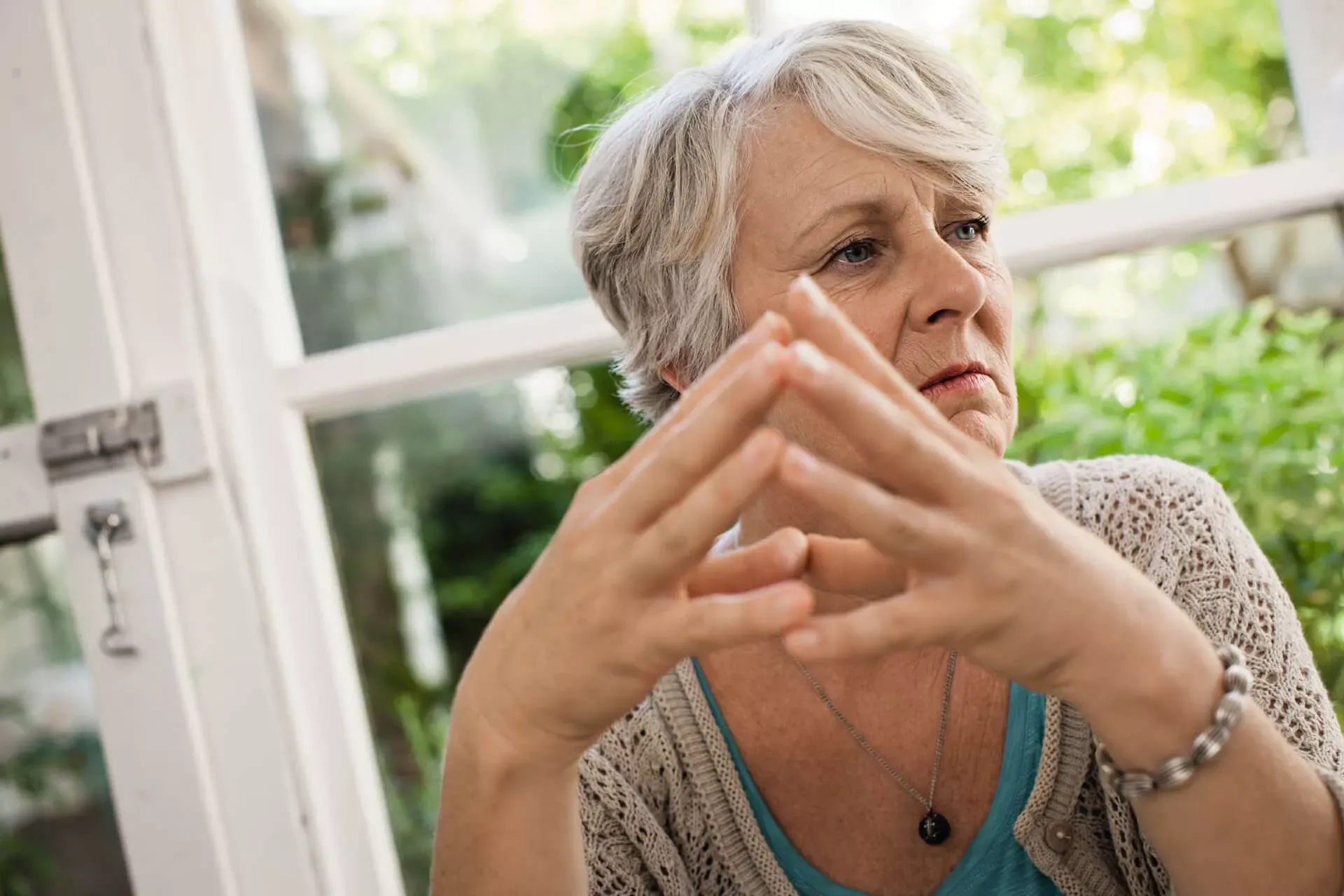 Porträt einer älteren Frau mit einem Fenster im Hintergrund. Sie legt mit kritischem Blick die Fingerspitzen aneinander. Porträt einer älteren Frau mit einem Fenster im Hintergrund. Sie legt mit kritischem Blick die Fingerspitzen aneinander.