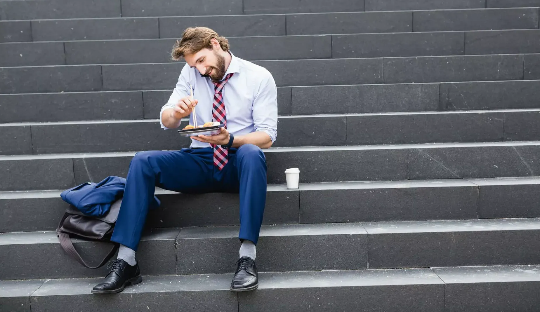 Mann in Businesskleidung sitzt auf Steintreppe mit Essen und Smartphone am Ohr. Mann in Businesskleidung sitzt auf Steintreppe mit Essen und Smartphone am Ohr.