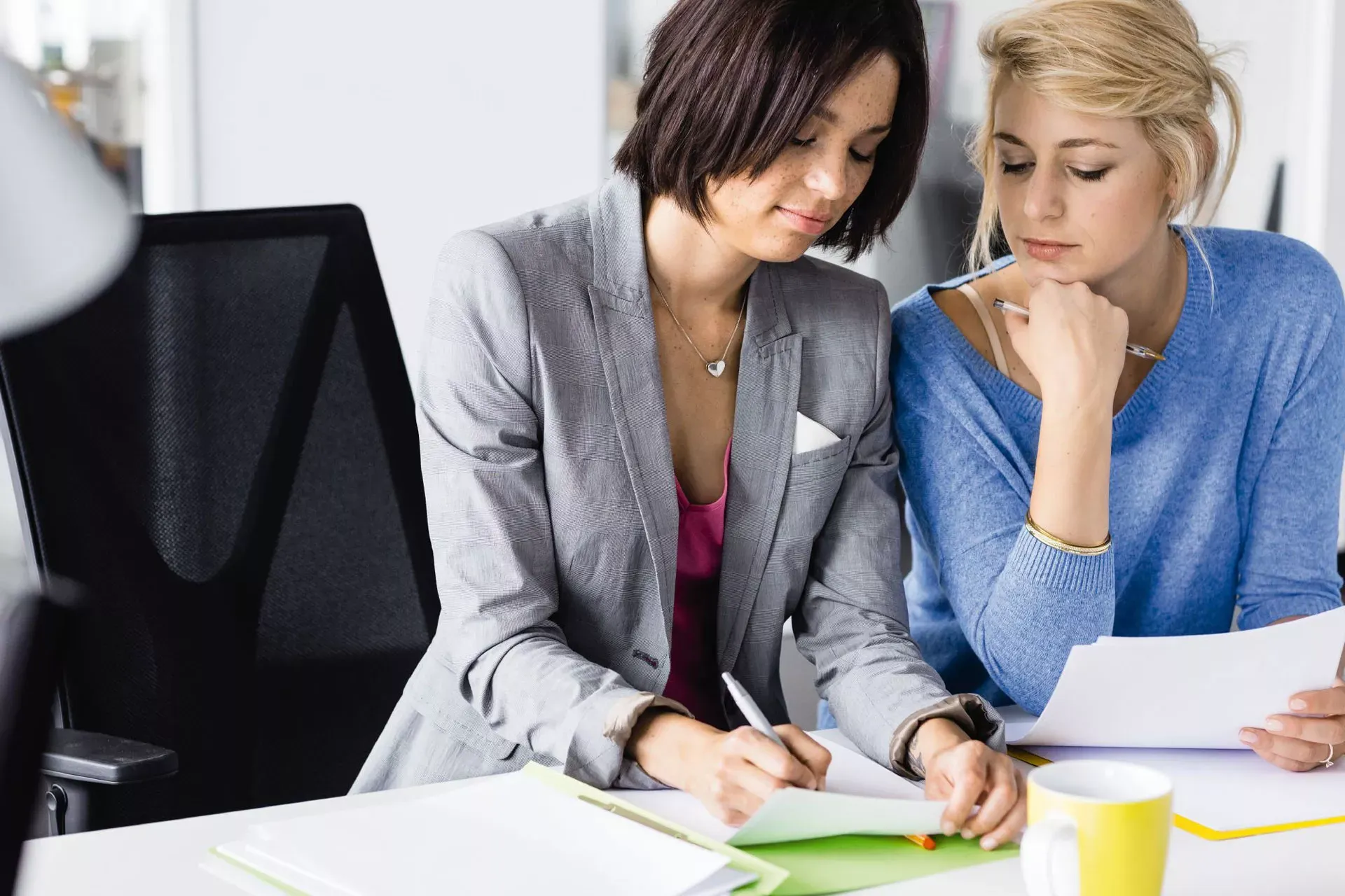 Zwei Kolleginnen im Büro sitzen zusammen an einem Schreibtisch und schauen in ein Dokument. Zwei Kolleginnen im Büro sitzen zusammen an einem Schreibtisch und schauen in ein Dokument.