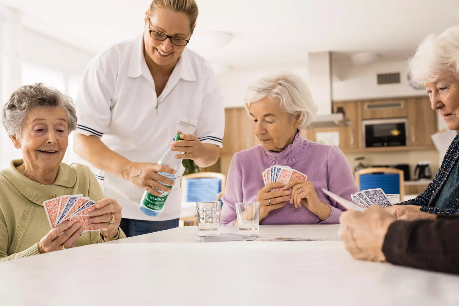 Seniorinnen im Pflegeheim spielen Karten, weibliche Pflegekraft schenkt ihnen Mineralwasser ein. Seniorinnen im Pflegeheim spielen Karten, weibliche Pflegekraft schenkt ihnen Mineralwasser ein.
