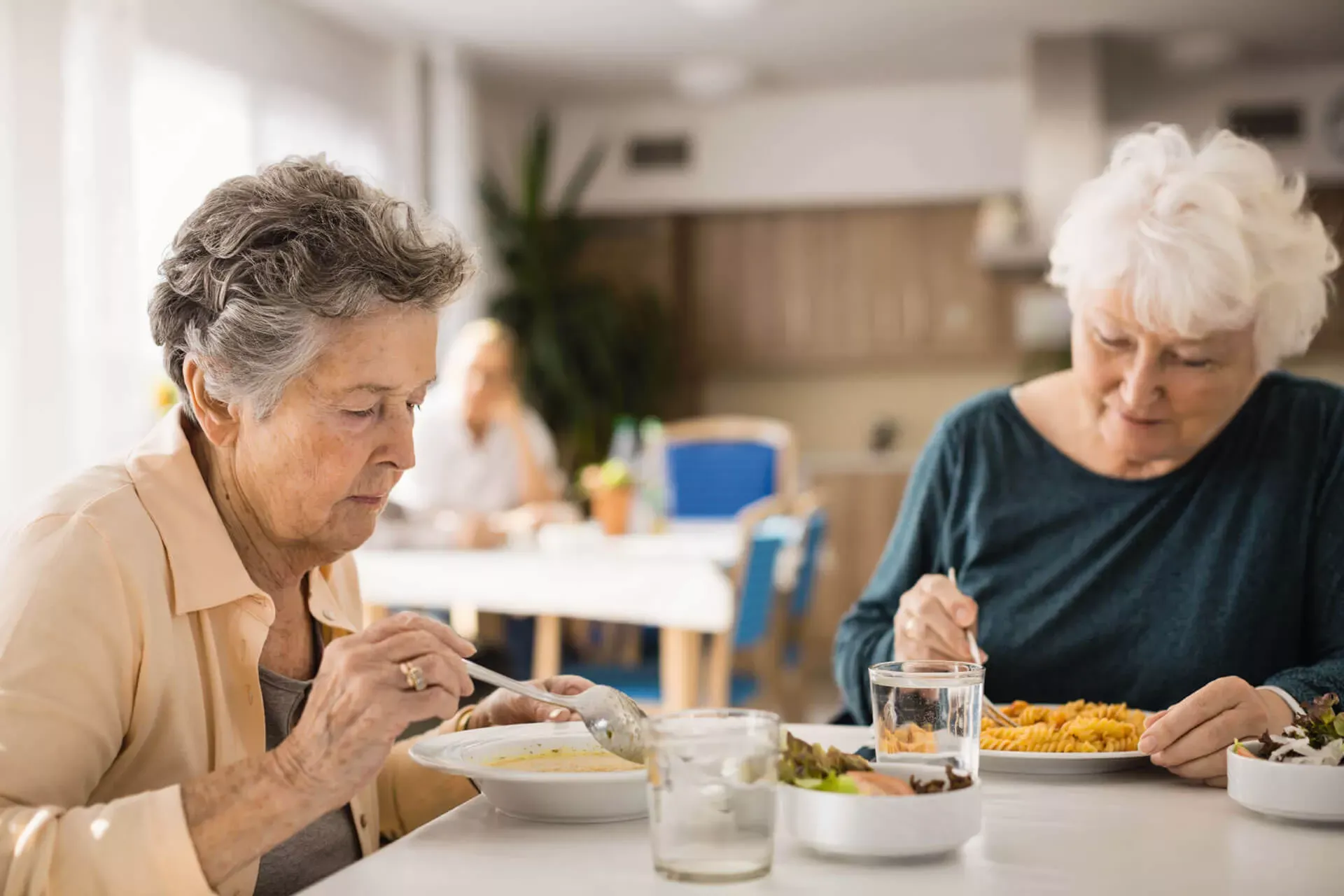 Zwei ältere Frauen sitzen an einem Tisch in einem Speisesaal und essen. Zwei ältere Frauen sitzen an einem Tisch in einem Speisesaal und essen.