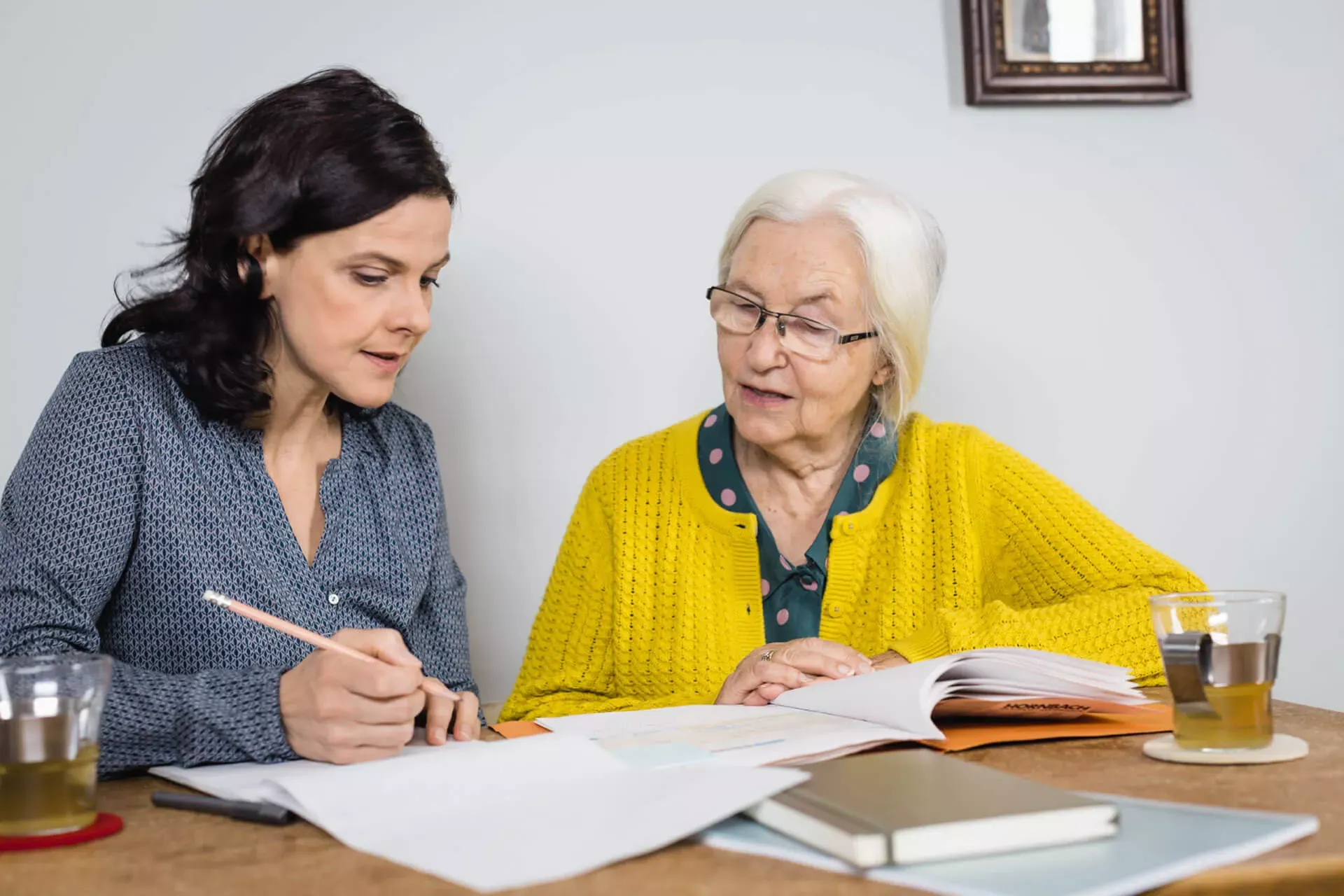 Seniorin und Frau im mittleren Alter sitzen an einem Tisch und prüfen zusammen Unterlagen. Seniorin und Frau im mittleren Alter sitzen an einem Tisch und prüfen zusammen Unterlagen.