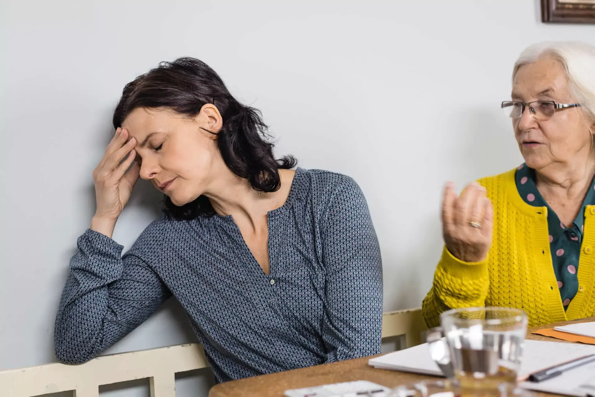 Pflegende Angehörige wendet sich genervt von Seniorin ab und legt die Finger an die Stirn. Pflegende Angehörige wendet sich genervt von Seniorin ab und legt die Finger an die Stirn.