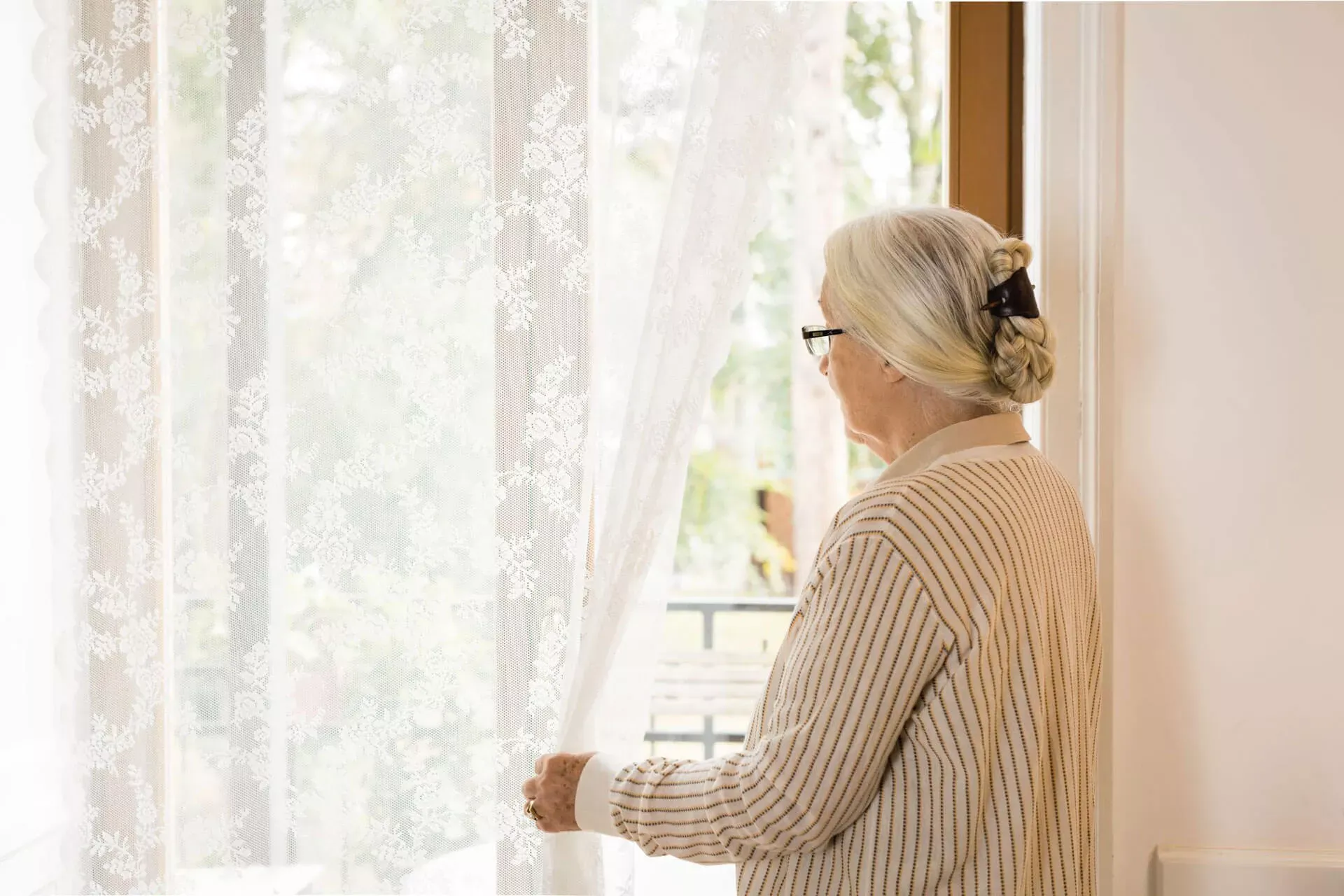 Seniorin mit Brille steht am Fenster, schiebt die Gardine zur Seite und schaut nach draußen. Seniorin mit Brille steht am Fenster, schiebt die Gardine zur Seite und schaut nach draußen.