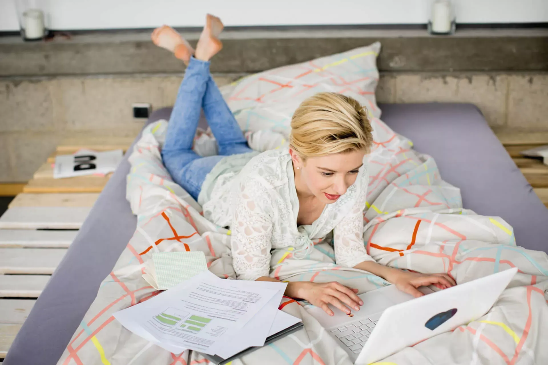 Eine junge Frau liegt auf einem Bett mit Unterlagen um sich herum und schaut in ihren Laptop. Eine junge Frau liegt auf einem Bett mit Unterlagen um sich herum und schaut in ihren Laptop.
