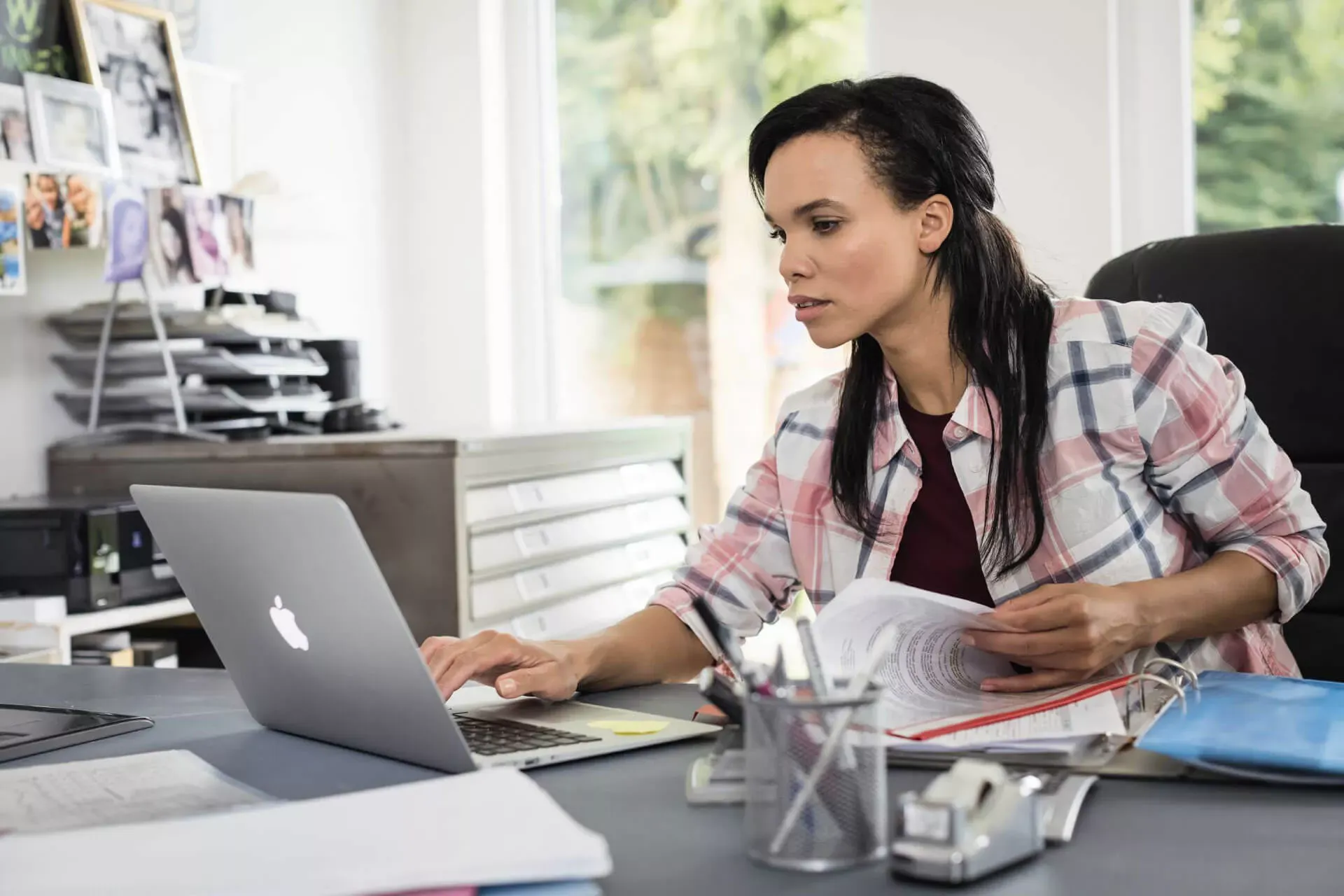 Junge Frau sitzt am Schreibtisch im Homeoffice, arbeitet an ihrem Laptop und blättert in einem Aktenordner. v Junge Frau sitzt am Schreibtisch im Homeoffice, arbeitet an ihrem Laptop und blättert in einem Aktenordner. v