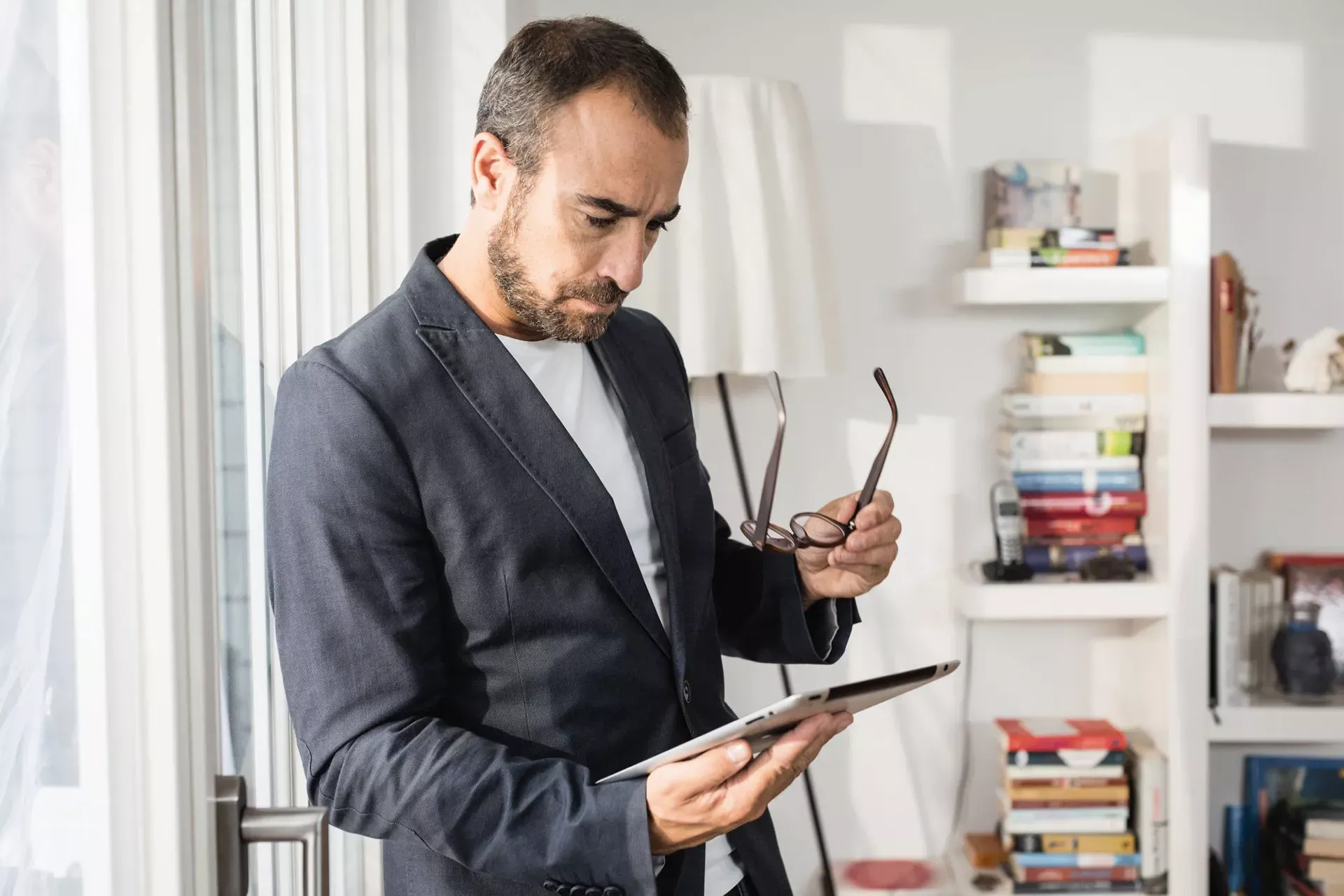 Mann mit Vollbart steht mit der Brille in der Hand in seinem Homeoffice und schaut auf ein Tablet. Mann mit Vollbart steht mit der Brille in der Hand in seinem Homeoffice und schaut auf ein Tablet.