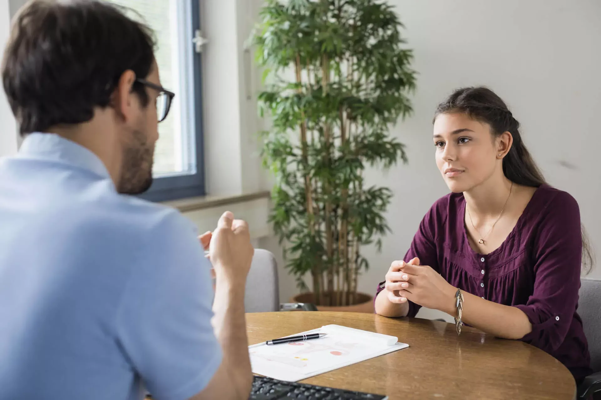 Jugendliche sitzt an einem runden Tisch im Gespräch mit einem Mann, auf dem Tisch liegt ein Dokument. Jugendliche sitzt an einem runden Tisch im Gespräch mit einem Mann, auf dem Tisch liegt ein Dokument.
