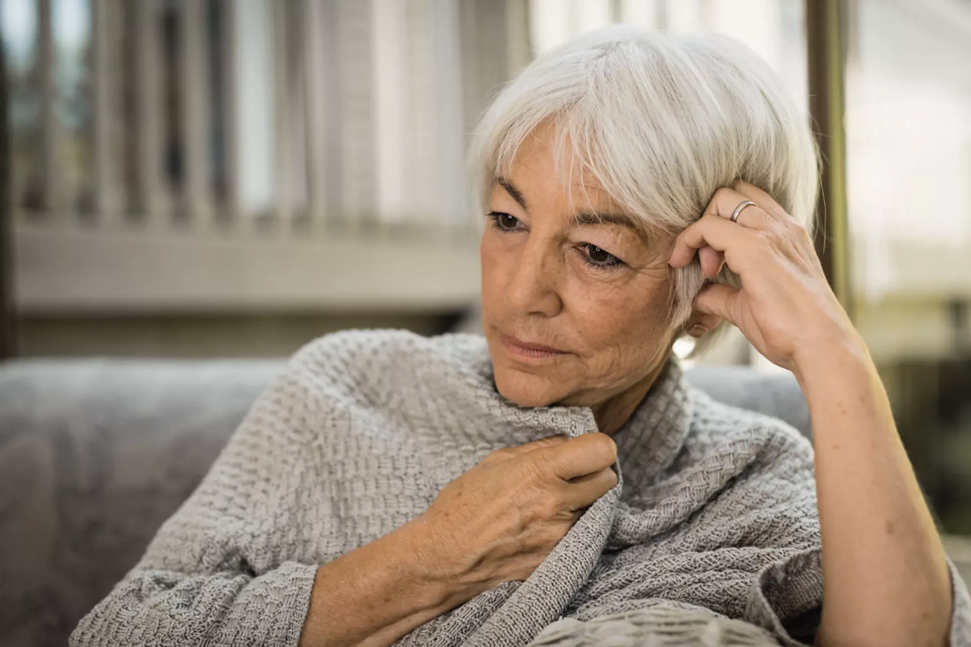 Seniorin sitzt nachdenklich auf einem Sofa, stützt ihren Kopf auf eine Hand und zieht eine Strickjacke enger um sich Seniorin sitzt nachdenklich auf einem Sofa, stützt ihren Kopf auf eine Hand und zieht eine Strickjacke enger um sich