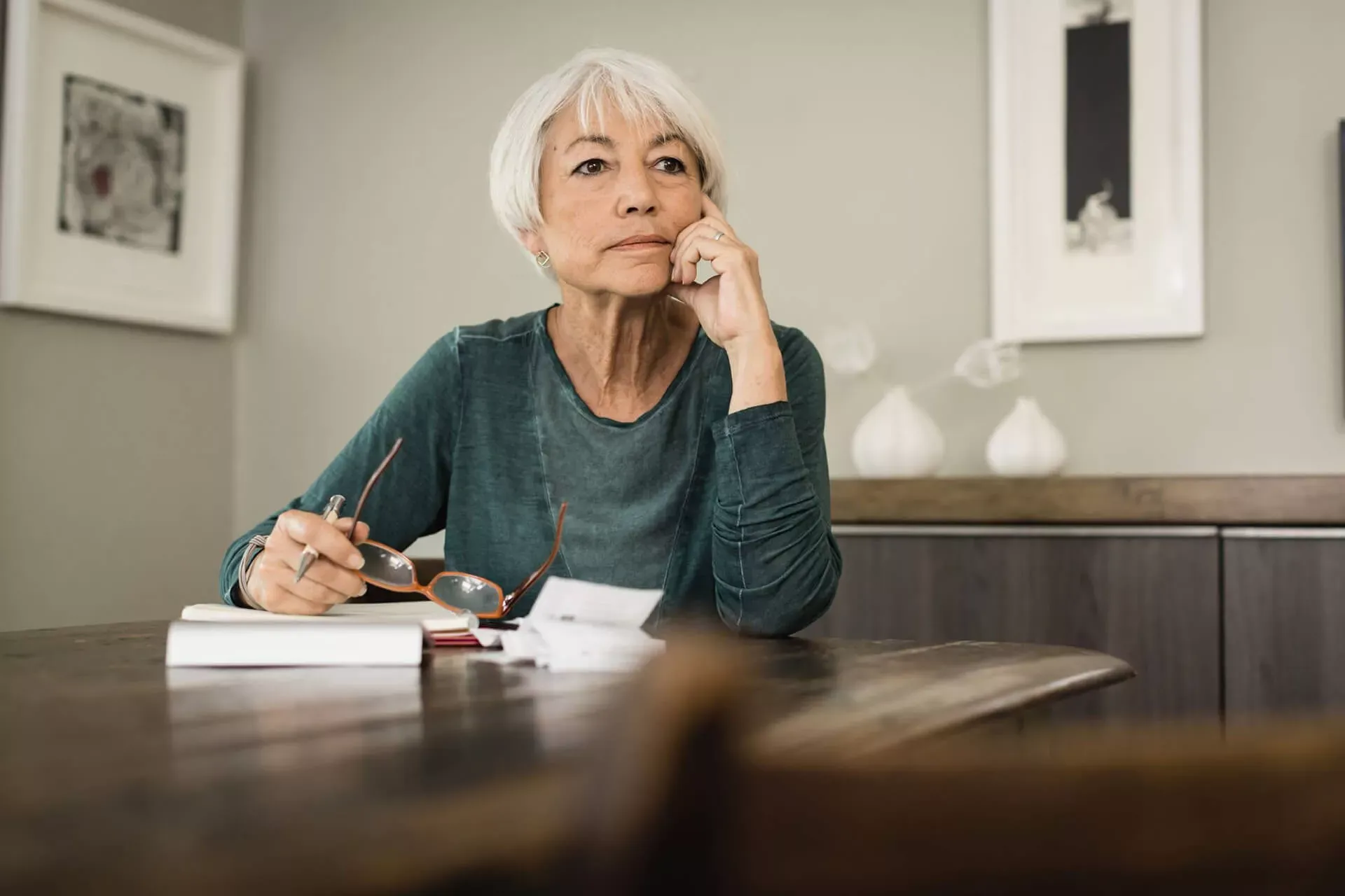 Seniorin sitzt im Wohnzimmer am Tisch über Belegen und Dokumenten und schaut nachdenklich in die Ferne. Seniorin sitzt im Wohnzimmer am Tisch über Belegen und Dokumenten und schaut nachdenklich in die Ferne.