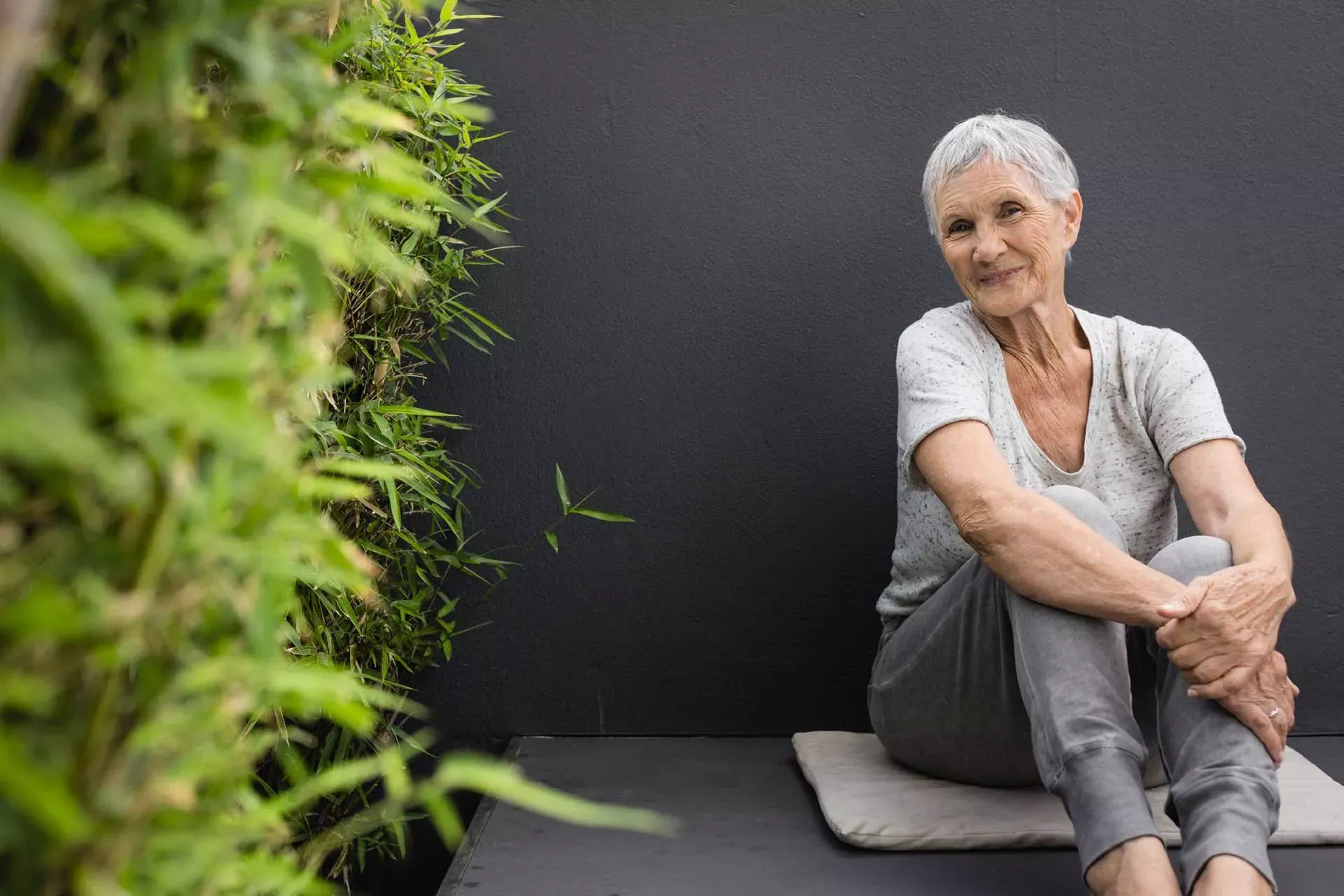 Lächelnde Seniorin sitzt mit angewinkelten Beinen auf dem Boden vor einer grauen Wand mit Bambuspflanzen an der Seite. Lächelnde Seniorin sitzt mit angewinkelten Beinen auf dem Boden vor einer grauen Wand mit Bambuspflanzen an der Seite.