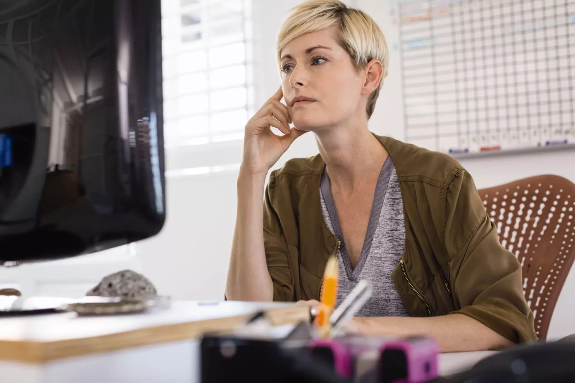 Junge Frau sitzt nachdenklich vor einem Computerbildschirm, im Hintergrund eine Personalplan-Tafel. Junge Frau sitzt nachdenklich vor einem Computerbildschirm, im Hintergrund eine Personalplan-Tafel.