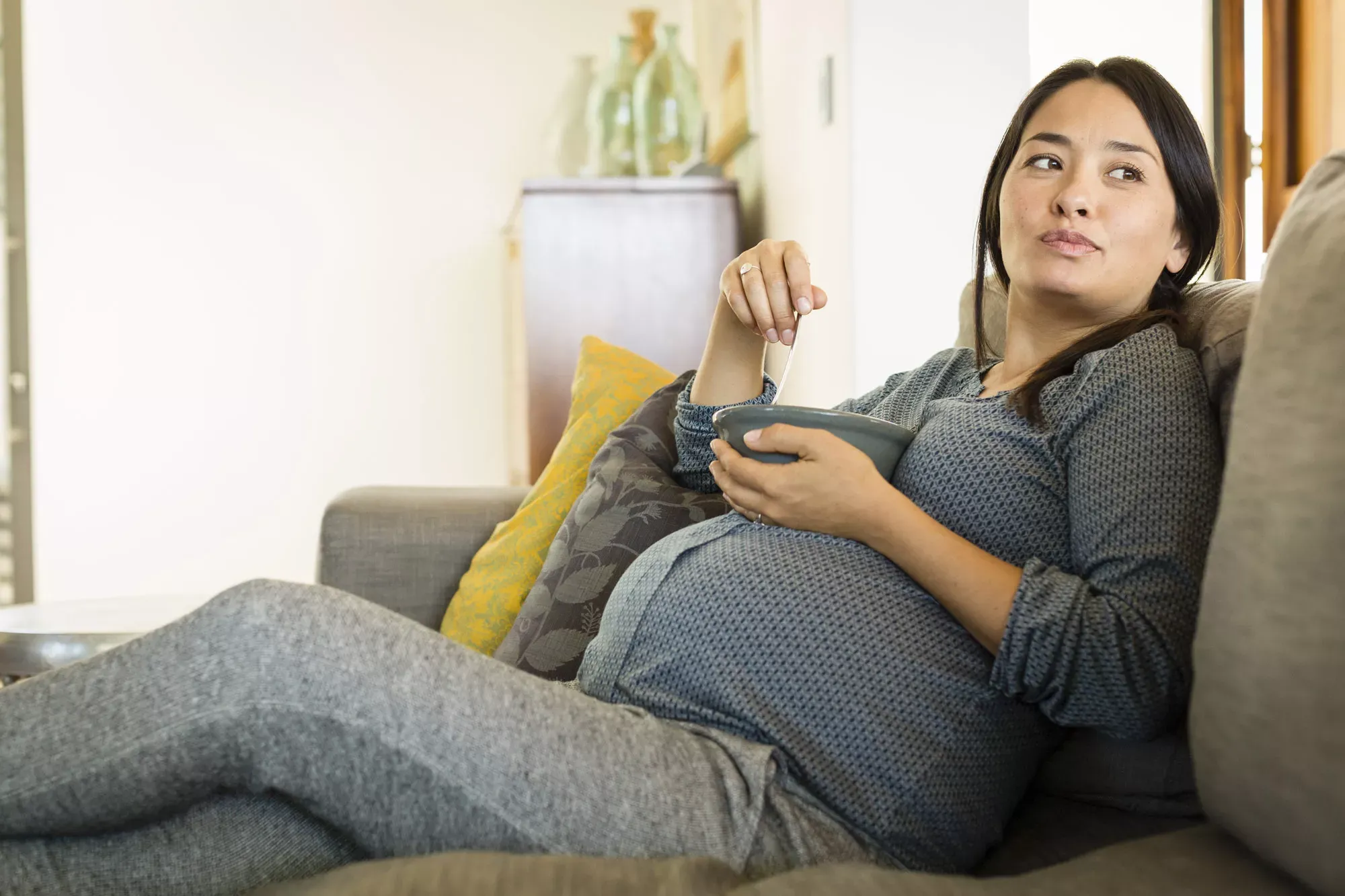 Junge schwangere Frau mit Migrationshintergrund sitzt auf einem Sofa und hält eine Schüssel in der Hand. Junge schwangere Frau mit Migrationshintergrund sitzt auf einem Sofa und hält eine Schüssel in der Hand.