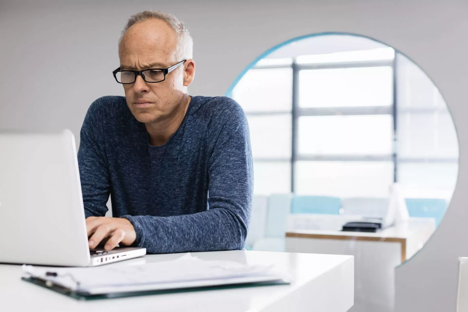 Ein Mann mittleren Alters mit Brille tippt mit gerunzelter Stirn auf der Tastatur seines Notebooks in einem Büro. Ein Mann mittleren Alters mit Brille tippt mit gerunzelter Stirn auf der Tastatur seines Notebooks in einem Büro.