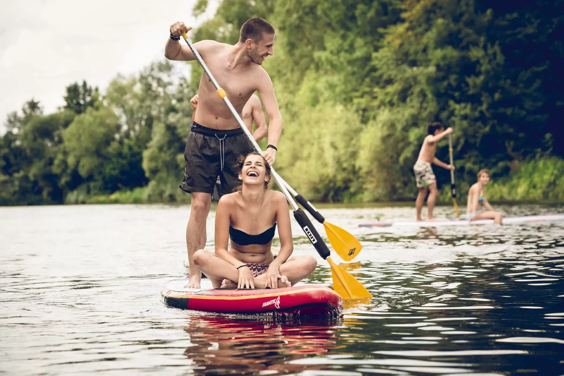 Junges Paar auf einem Stand-up-Paddling-Board, der Mann paddelt im Stehen, die Frau sitzt vorne im Schneidersitz und lacht. Junges Paar auf einem Stand-up-Paddling-Board, der Mann paddelt im Stehen, die Frau sitzt vorne im Schneidersitz und lacht.