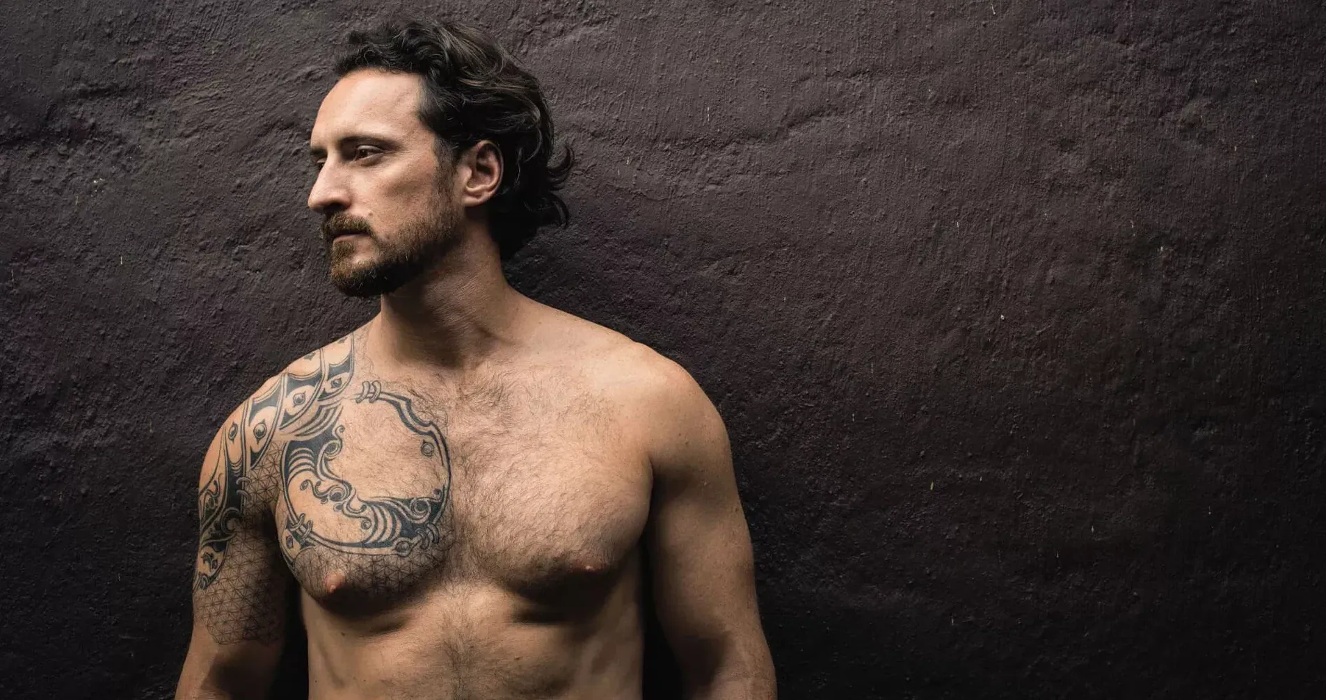 Mann mit nacktem Oberkörper und Tattoos steht vor einer dunklen Wand. Mann mit nacktem Oberkörper und Tattoos steht vor einer dunklen Wand.